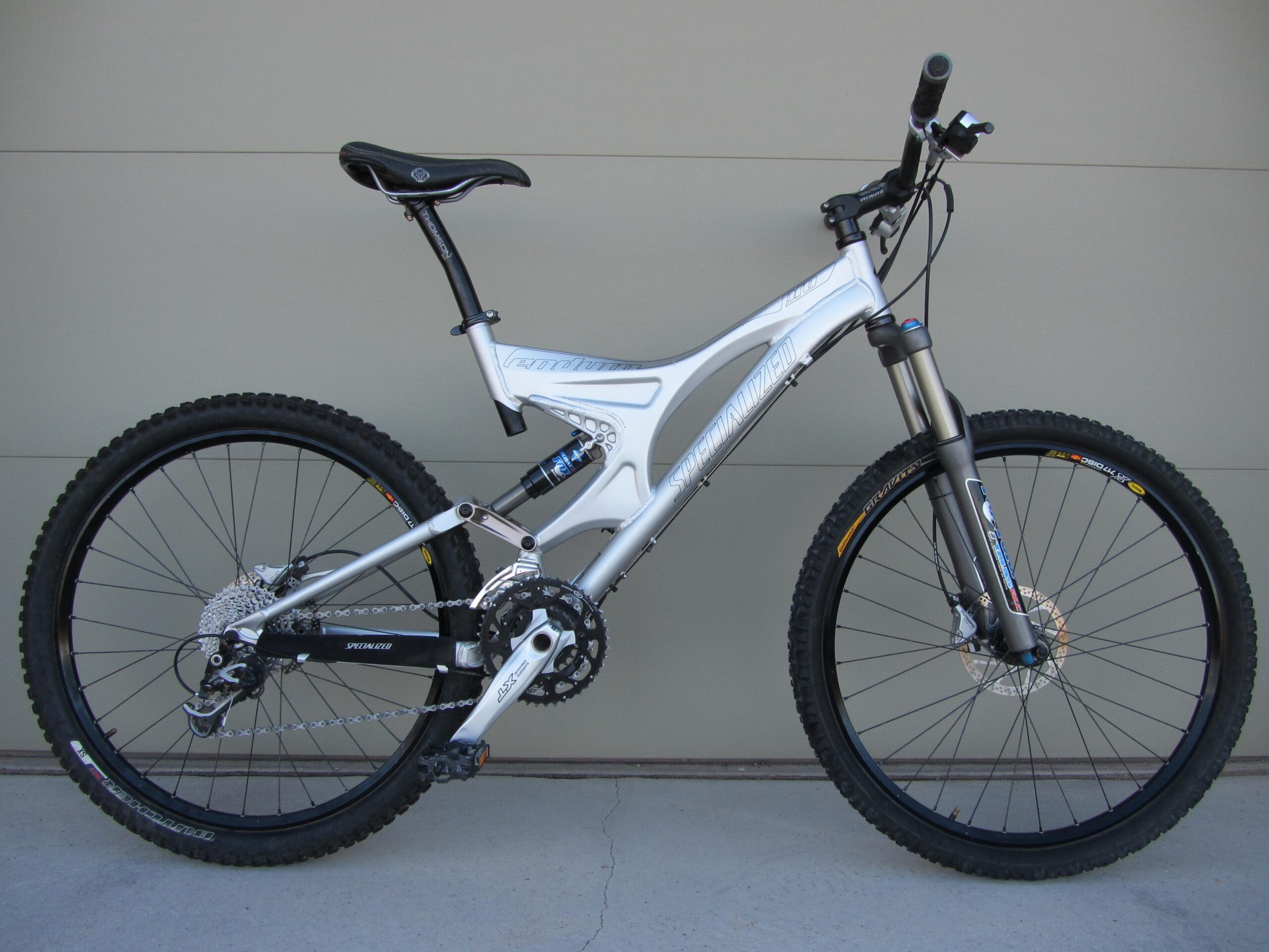 A side view of a Specialized mountain bike featuring a silver frame, front suspension fork, and knobby tires suitable for off-road riding. The bike is positioned against a plain background, highlighting its design and components, including gears and brakes.