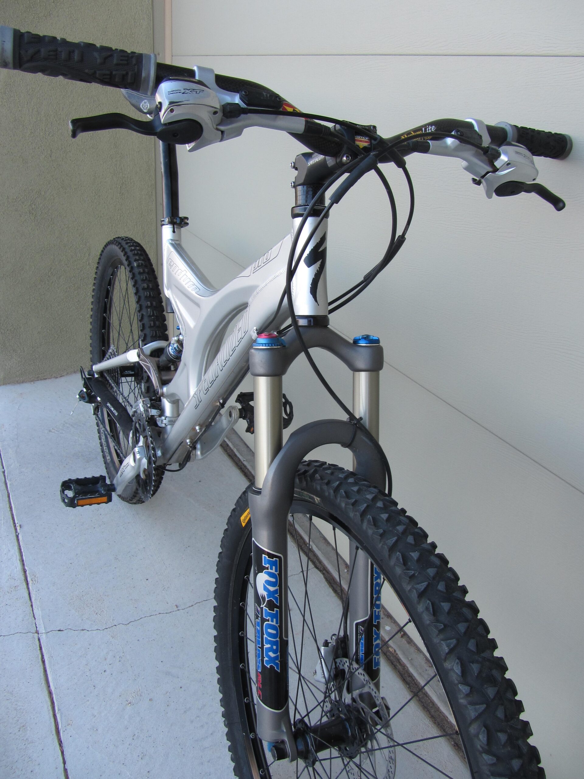 A close-up view of a modern mountain bike featuring a silver frame, dual suspension setup, and prominent front fork with branding. The handlebars are equipped with grips and gear shifters, and the bike is positioned against a light-colored wall with a smooth concrete surface beneath it.