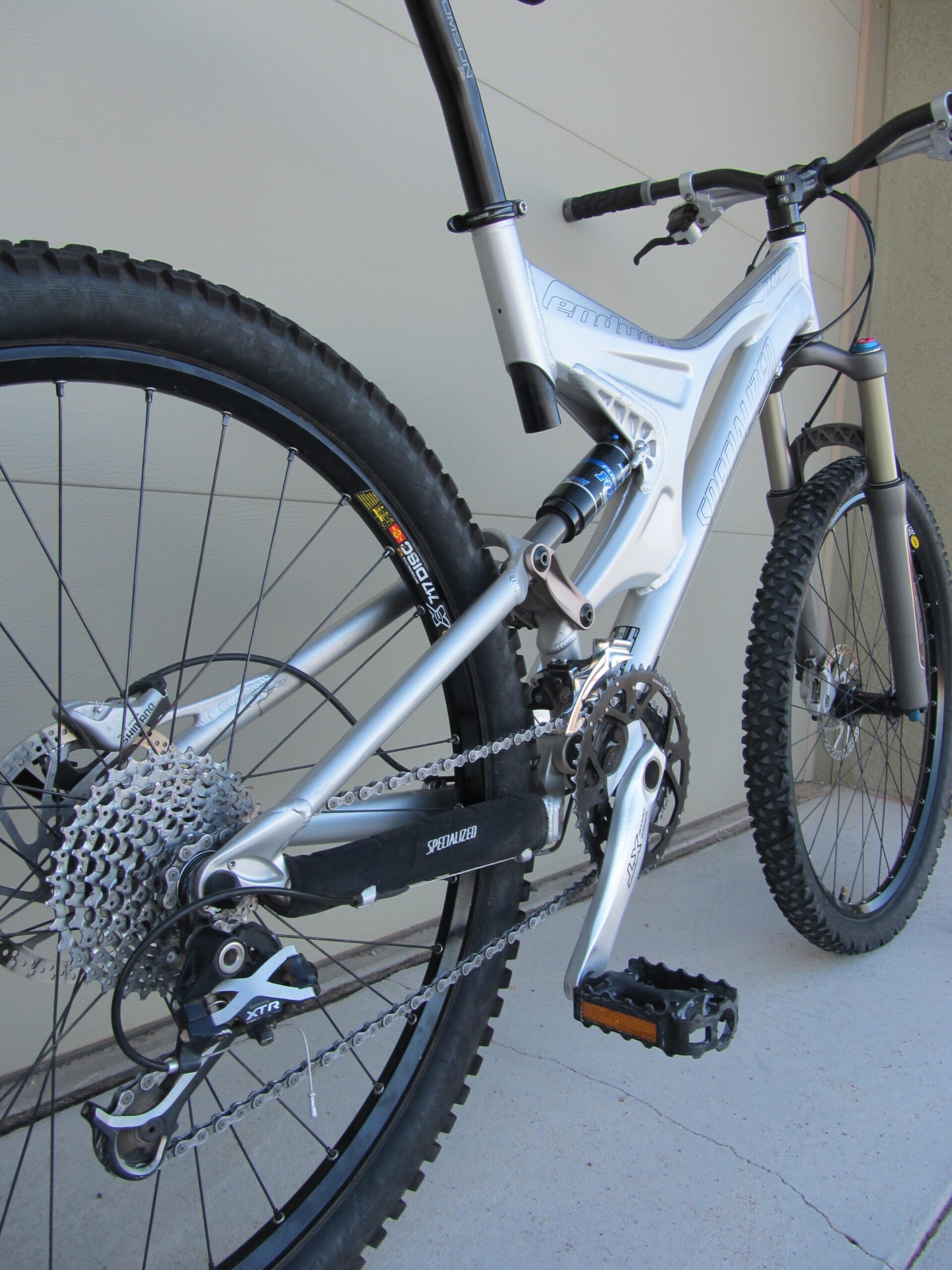 Close-up of a silver mountain bike, featuring detailed components such as a rear derailleur, cassette, and pedal. The bike is positioned against a plain background, highlighting its rugged tires and suspension system.