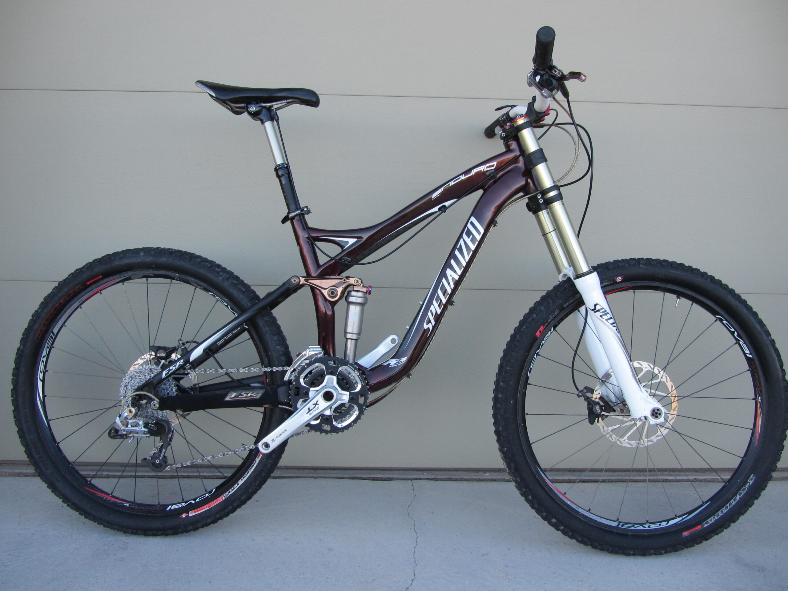 Alt text: A Specialized mountain bike featuring a dark brown frame, front suspension forks, and wide tires, displayed against a light-colored wall. The bike is equipped with hydraulic disc brakes and a rear shock absorber, showcasing its durable construction and design for off-road cycling.