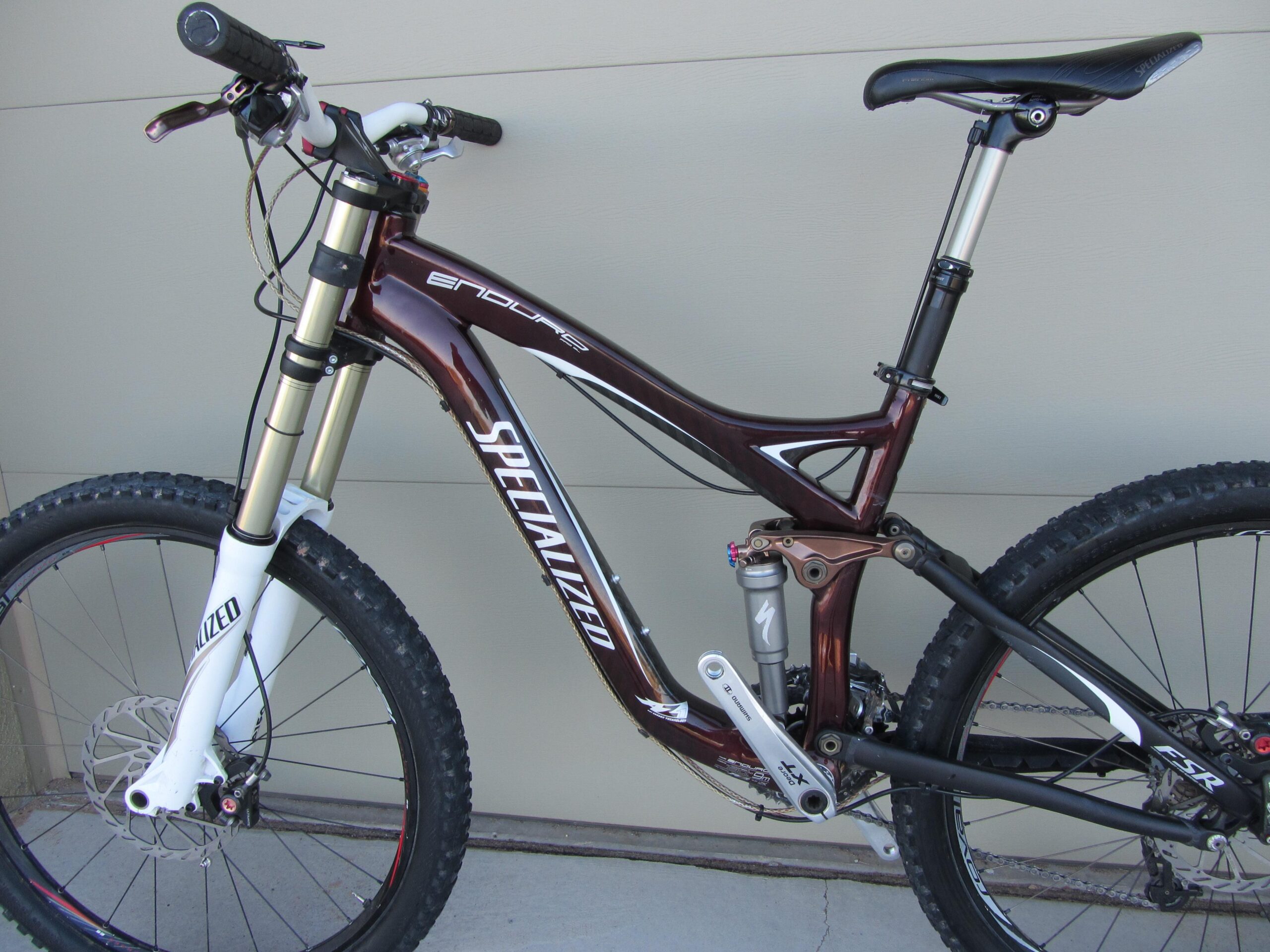 Alt text: Close-up view of a Specialized Enduro mountain bike featuring a maroon and white frame, front suspension fork, and knobby tires, set against a neutral background.