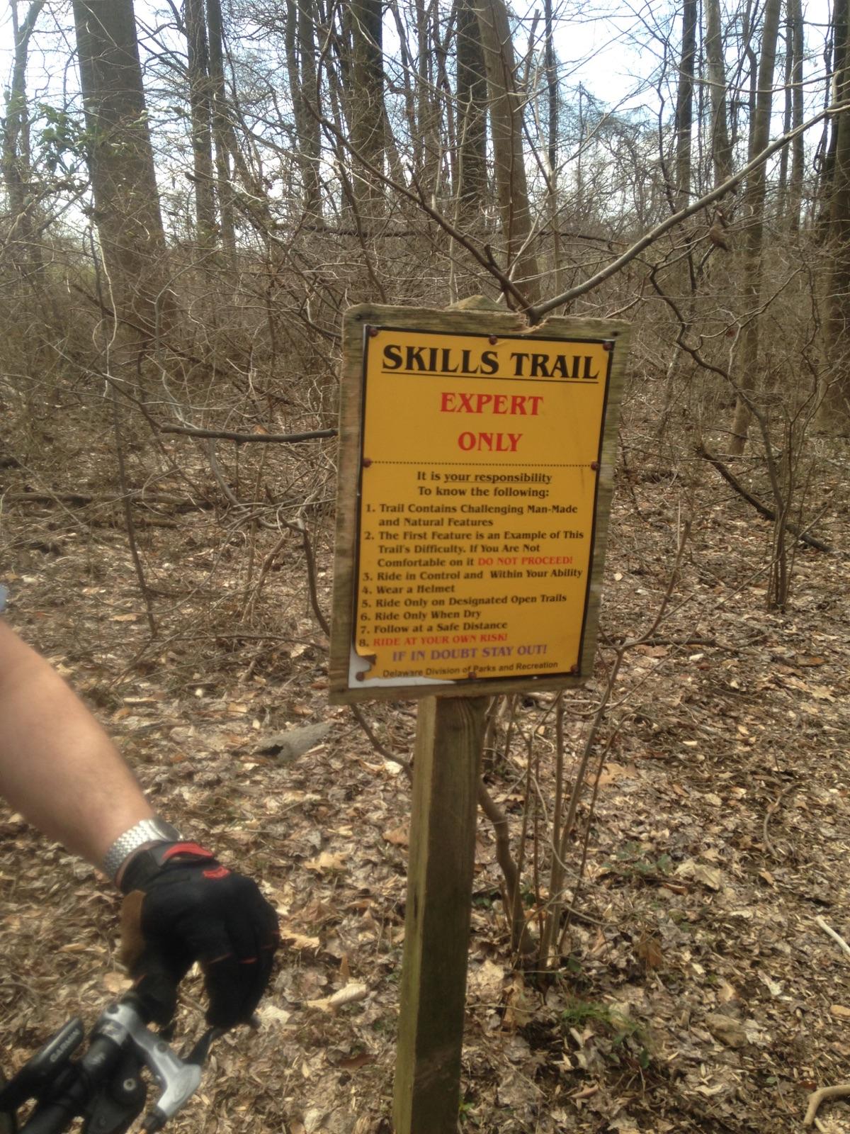 Sign indicating the "Skills Trail" designated for "Expert Only" riders, surrounded by a wooded area. The sign includes safety and usage guidelines for the trail, emphasizing the risks and responsibilities of riders. A person wearing cycling gloves and holding a bike's handlebars is partially visible in the foreground. White Clay Creek mountain bike trail.