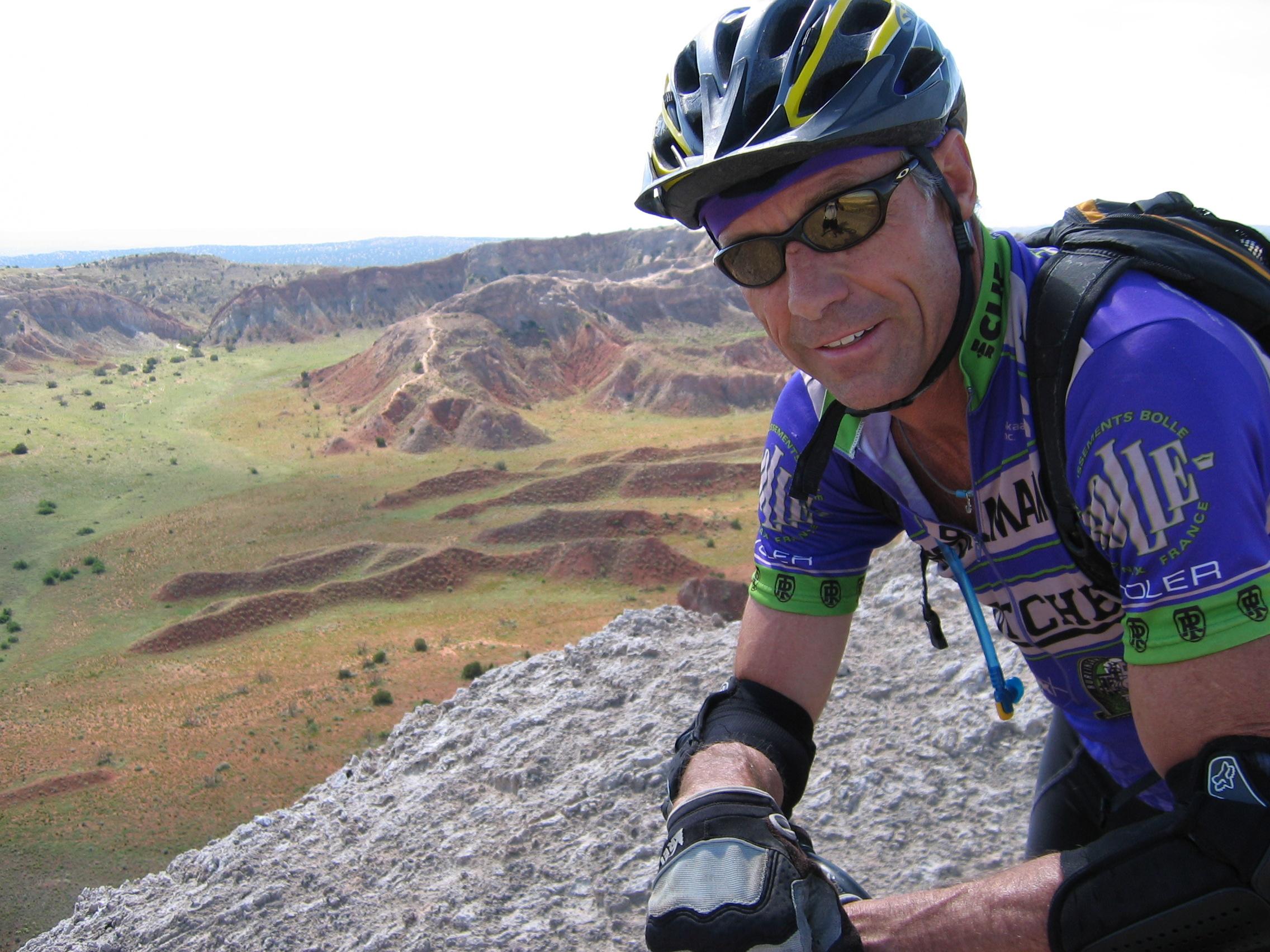 A cyclist wearing a helmet and sunglasses poses on a rocky outcrop, with a scenic view of rolling hills and colorful terrain in the background. The cyclist is dressed in a blue and green cycling jersey and gloves, showcasing a smile as they enjoy the outdoors.