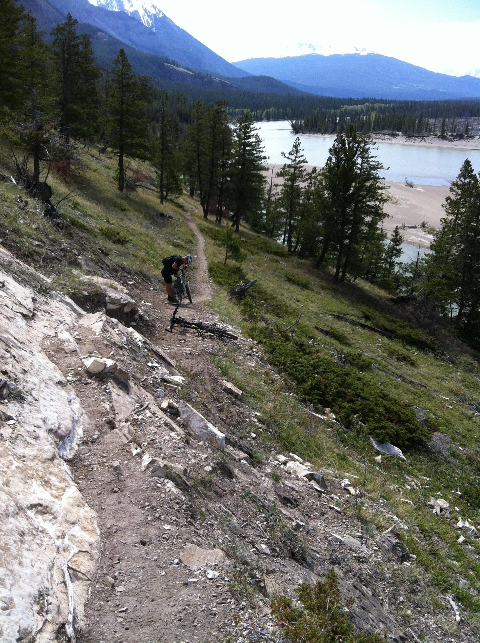 A person crouches on a hillside trail, adjusting a mountain bike while surrounded by trees and a view of a river and sandy area in the background. Snow-capped mountains are visible in the distance under a partly cloudy sky. The Overlander mountain bike trail.
