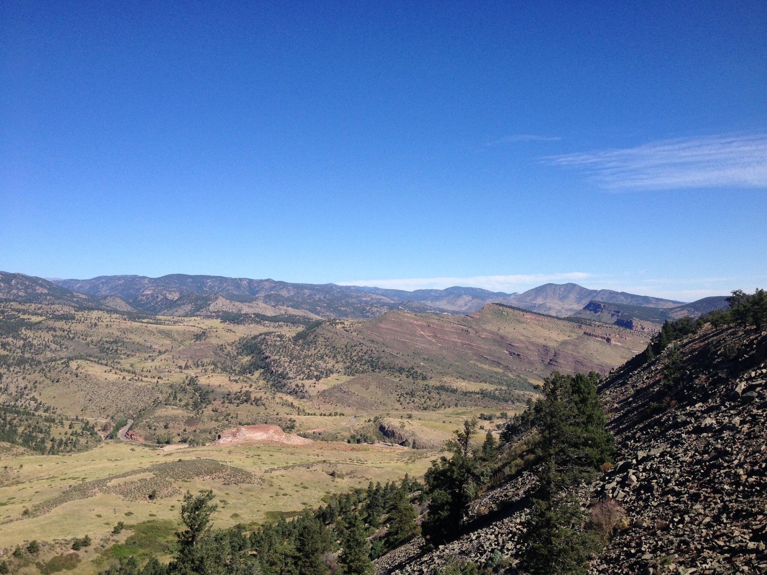 A panoramic view of a mountainous landscape under a clear blue sky, featuring rolling hills, scattered trees, and rocky terrain. The scene captures the beauty of nature with a blend of greenery and earthy tones, showcasing distant mountain ranges. Heil Valley Ranch mountain bike trail.
