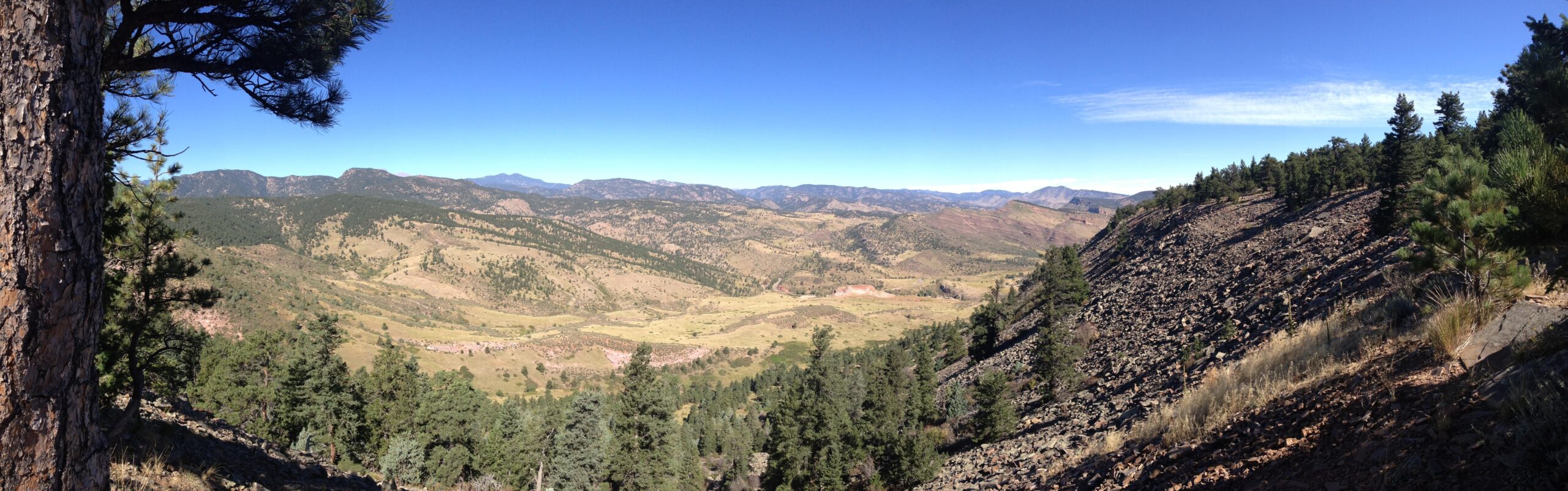 A panoramic view of a mountainous landscape featuring rolling hills and valleys, with patches of green forests interspersed. The sky is clear and blue, showcasing a few wispy clouds, while rocky terrain is visible in the foreground. Heil Valley Ranch mountain bike trail.