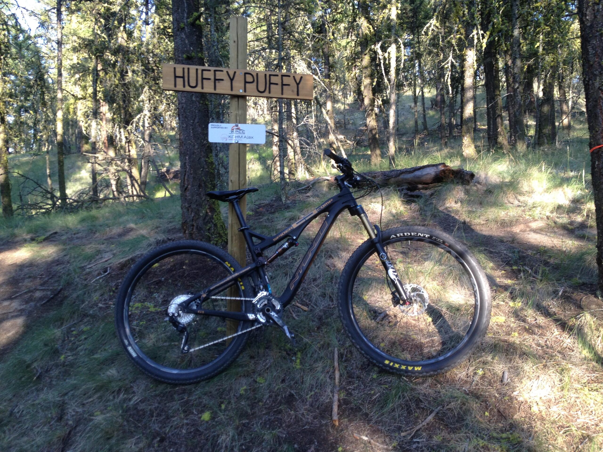 A black mountain bike is leaning against a wooden sign labeled "Huffy Puffy" in a forested area. The surroundings include tall trees and patches of grass, indicating a popular biking trail. Pineview mountain bike trail.