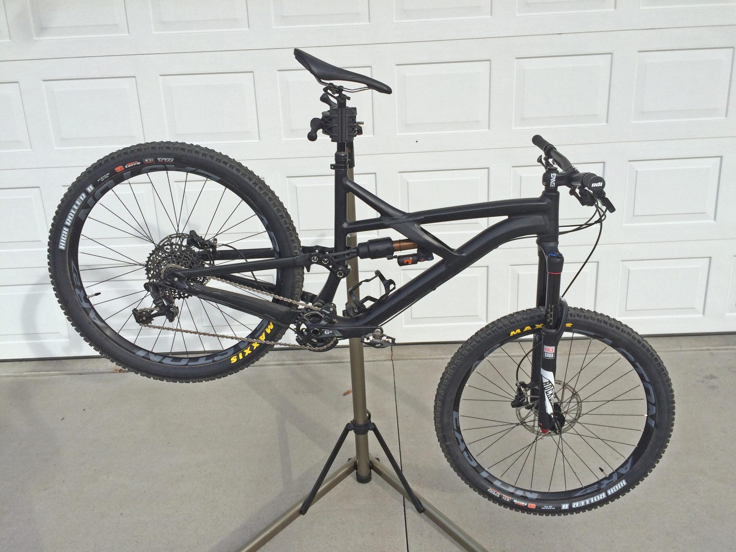 Specialized Enduro Comp 650b: A black mountain bike suspended in a repair stand, showcasing its frame and wheels. The rear wheel is slightly lifted off the ground, revealing the drivetrain. The bike features thick, knobby tires and a modern design suitable for off-road riding. In the background, a white garage door can be seen.