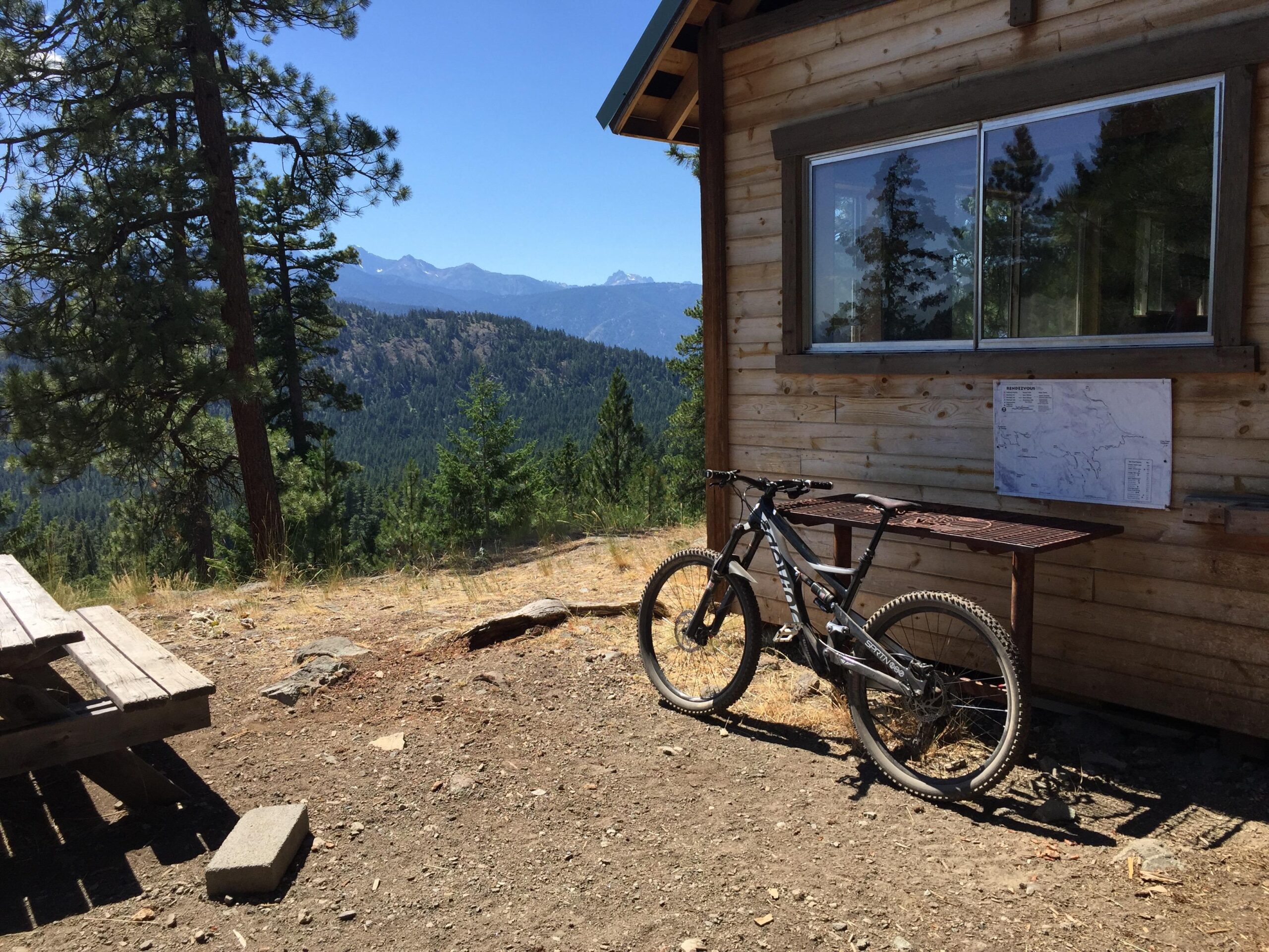 Devinci Spartan: A mountain bike rests against a wooden table outside a cabin, with a scenic view of mountains and trees in the background. The sky is clear and blue, and a map is mounted on the cabin wall. A wooden picnic table is visible to the left.