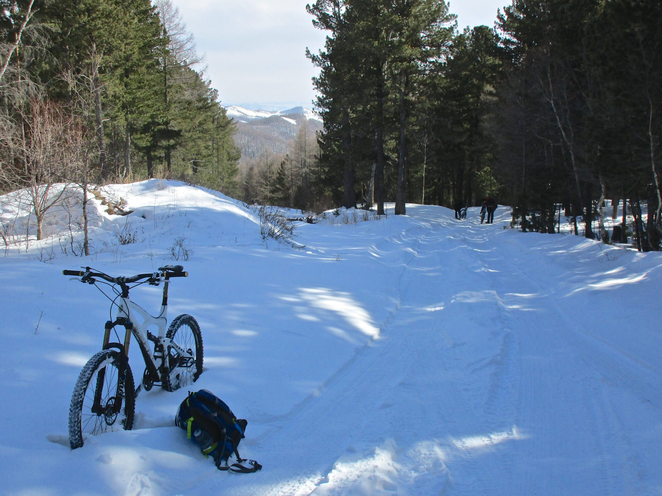 A snowy outdoor trail surrounded by tall pine trees, with a mountain bike resting in the foreground next to a backpack. In the distance, two people are walking along the path, enjoying the winter scenery. Soft sunlight illuminates the landscape, creating a serene winter atmosphere. Shargamorit Khandgait Hills East mountain bike trail.