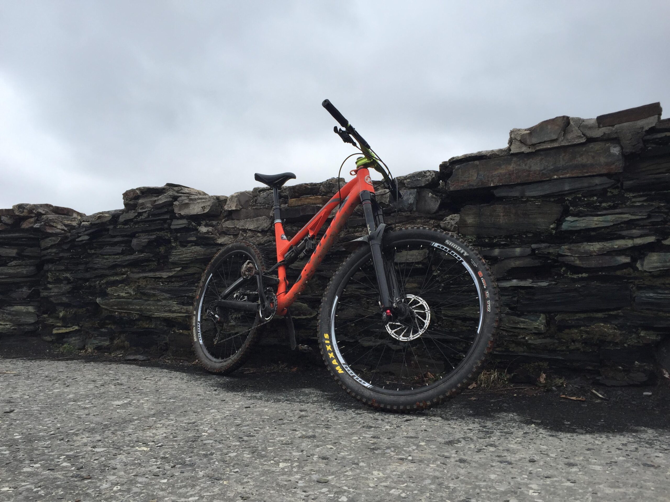 Kona Process 153 DL: A mountain bike in vibrant orange color resting against a stone wall. The scene is set on a gray, overcast day, with a paved pathway visible in the foreground.