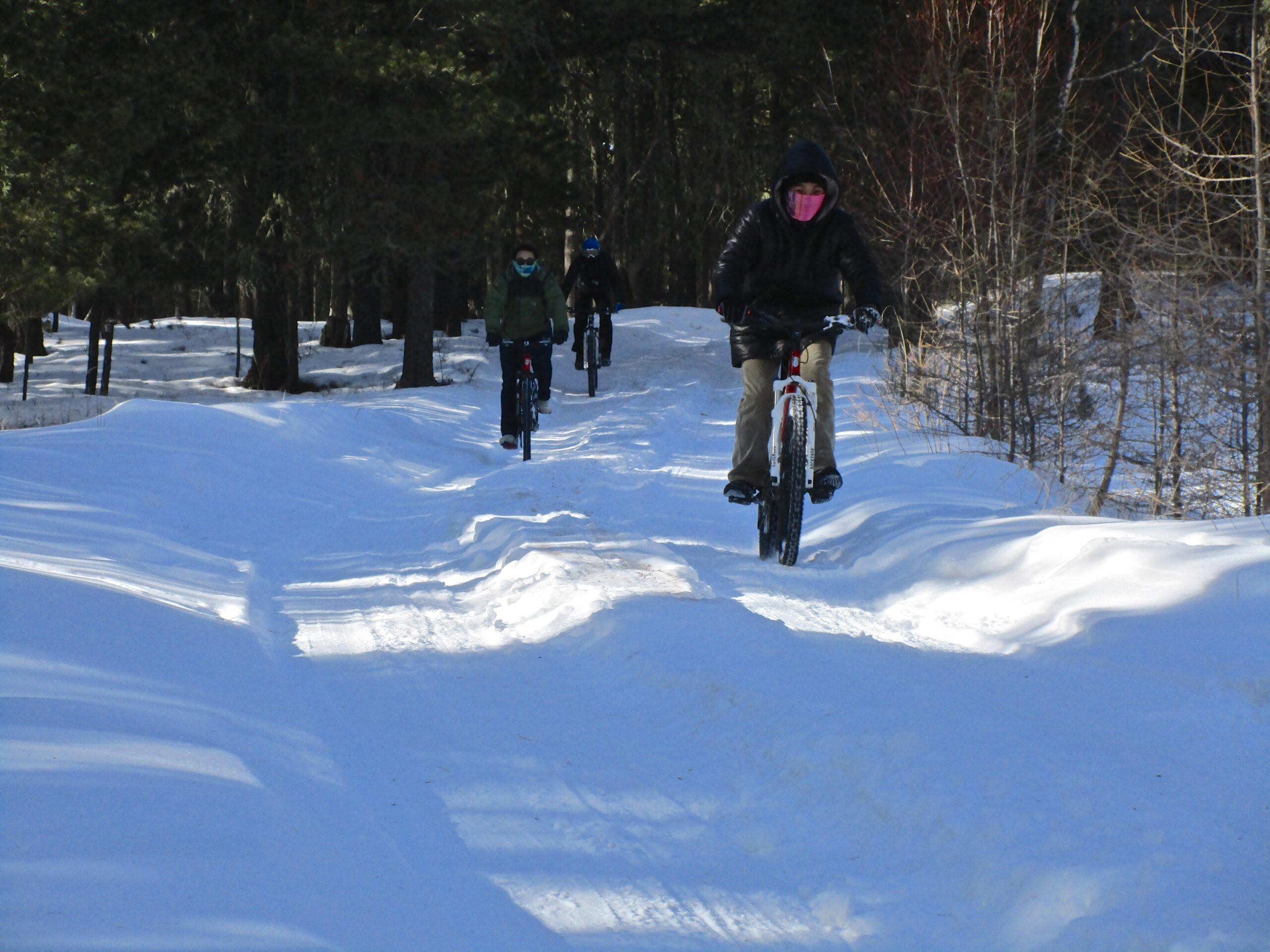 Three people are riding bicycles on a snowy trail surrounded by trees. The first rider, in the foreground, is wearing a black jacket and a mask, while the others are further back, dressed in winter clothing. The path shows signs of use, with patches of snow and clear tire tracks. The scene depicts a bright, wintry day. Shargamorit Khandgait Hills East mountain bike trail.