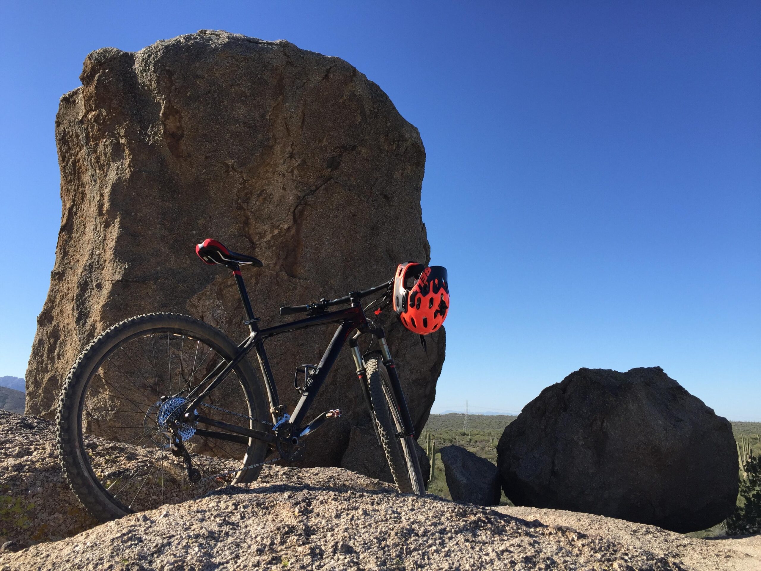 Specialized Carve Expert 29er: A mountain bike with a red and black helmet rests against a large rock formation in a desert landscape under a clear blue sky.