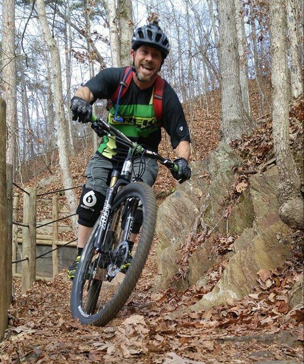 A mountain biker riding on a rocky trail surrounded by trees, smiling as he navigates the path. He is wearing a black helmet, a green and black cycling jersey, and knee pads, with a backpack secured to his back. The ground is covered with dry leaves, and the surrounding woods are in the background. Blankets Creek mountain bike trail.