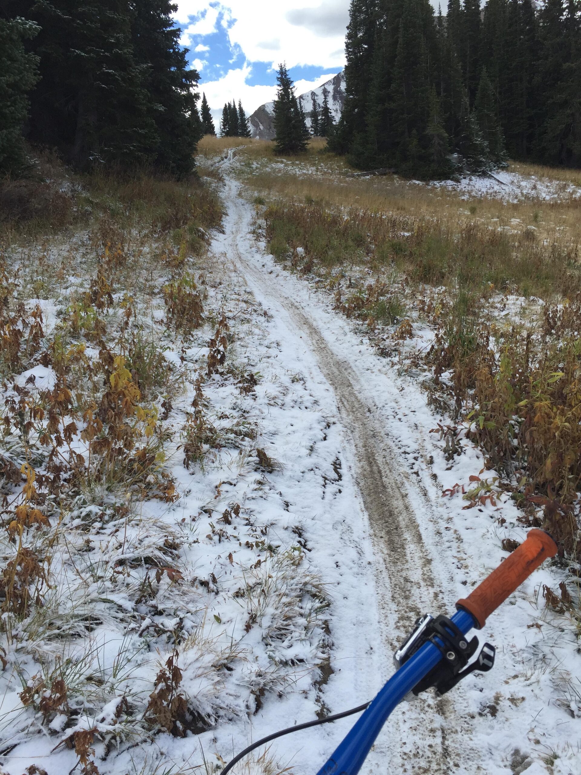 A snow-covered bike trail winding through a mountain landscape, with autumn foliage on either side and a cyclist's handlebars visible in the foreground. The sky is partly cloudy, and snow is lightly dusting the ground. Trail 401 mountain bike trail.