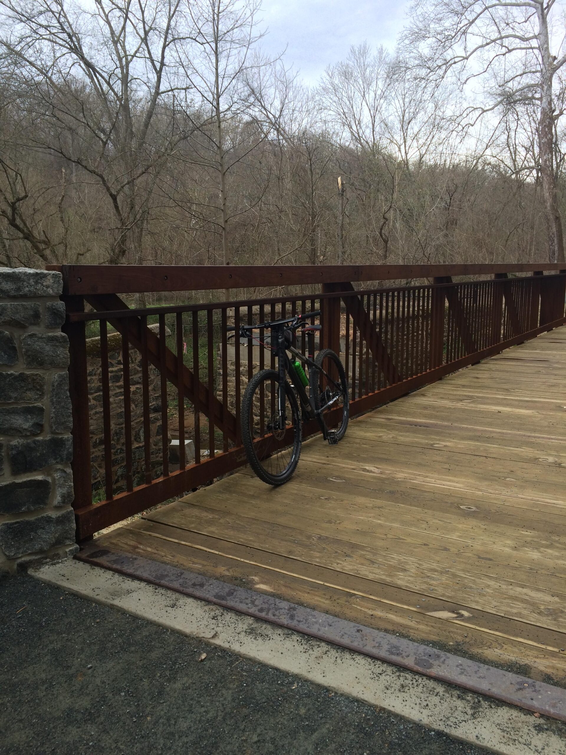 A mountain bike leaning against a wooden rail on a bridge, surrounded by bare trees and a wooded landscape under a cloudy sky. Brandywine State Park mountain bike trail.