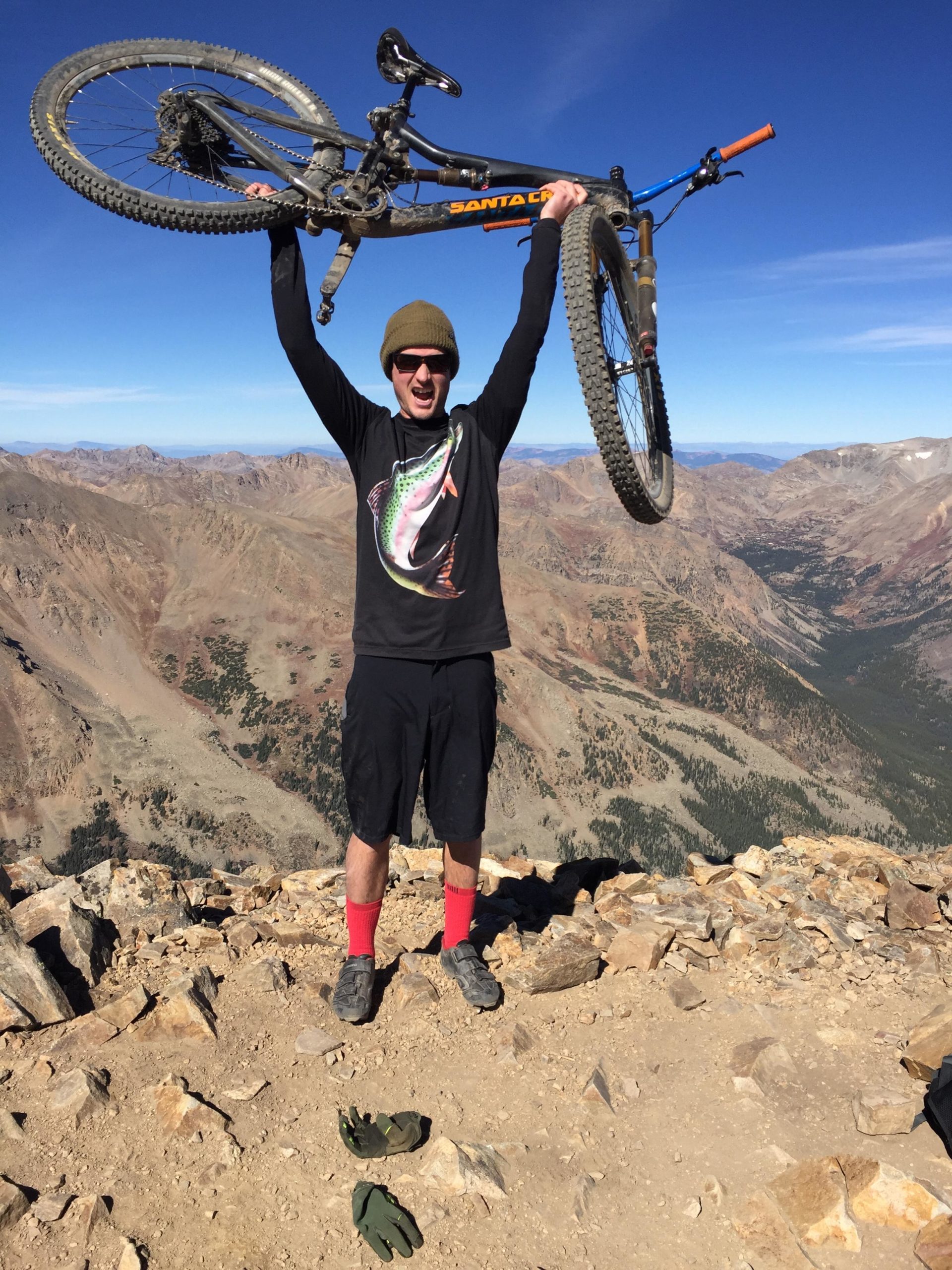 A person standing triumphantly on a mountain peak, holding a mountain bike overhead. The individual is wearing a black long-sleeve shirt with a colorful fish design, shorts, and bright red socks. The background features a panoramic view of rugged mountain terrain under a clear blue sky. Two pairs of gloves are placed on the ground nearby. Trail 401 mountain bike trail.