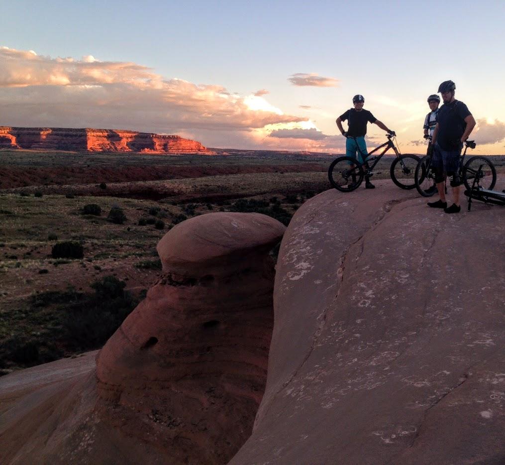 Three mountain bikers stand on a rocky outcrop overlooking a vast desert landscape at sunset. The sky is filled with soft clouds, and the distant cliffs are illuminated by a warm golden light. The terrain features a mix of red rock formations and green vegetation, creating a striking natural scene. Bartlett Wash mountain bike trail.