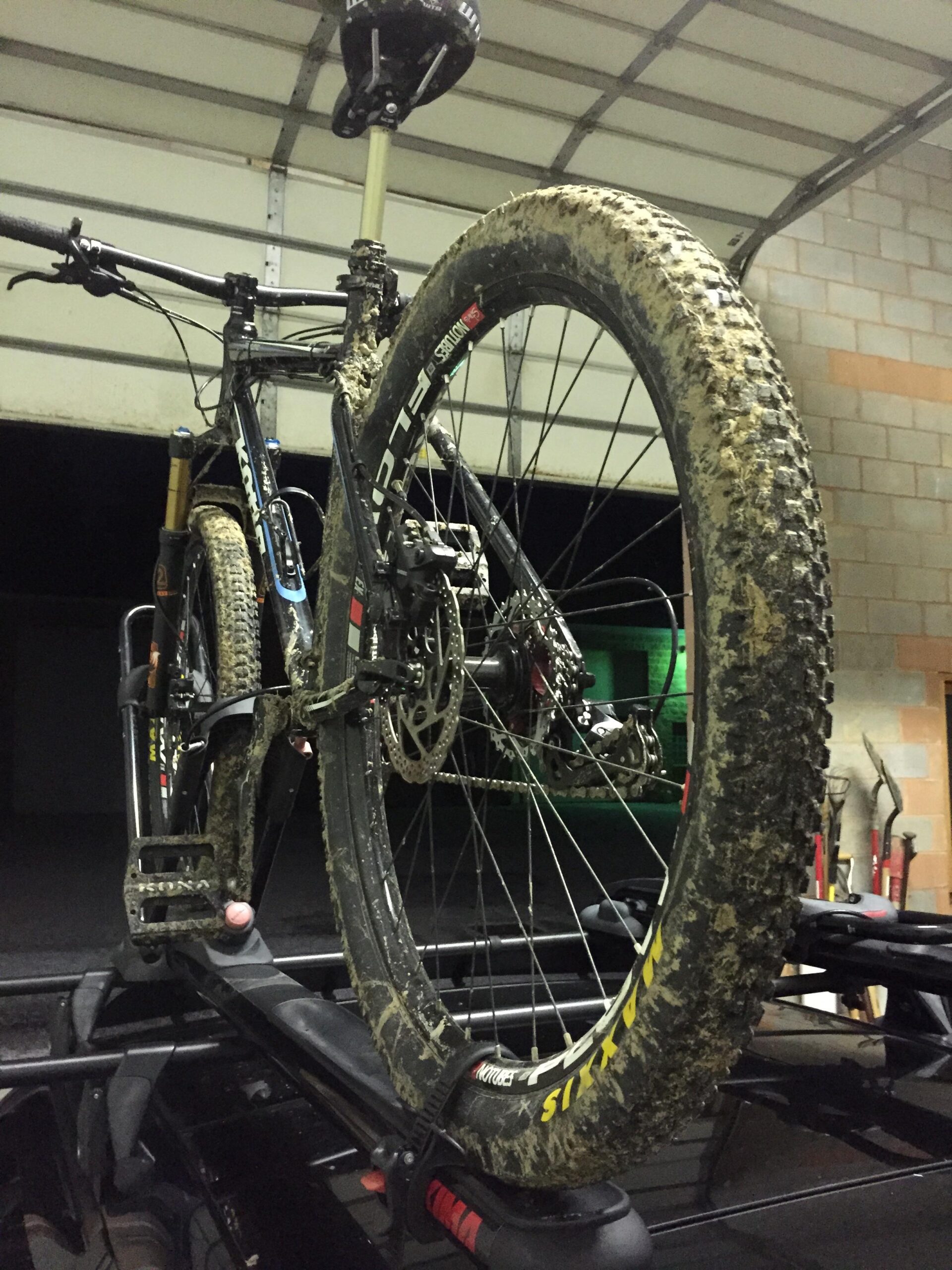 Maxxis Ardent Race: A mountain bike with muddy tires is secured on a roof rack, inside a garage with dim lighting. The bike is positioned upright, showcasing its suspension, handlebars, and rear wheel covered in mud, indicating recent use on a rugged terrain. The garage features a simple background with a few tools visible.