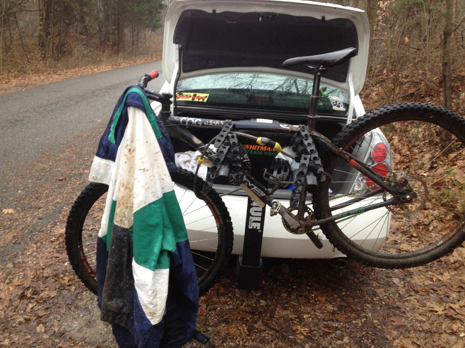 A mountain bike hanging on the back of a car with its rear wheel resting against the vehicle. A partially muddy jacket is draped over the bike frame. The car's trunk is open, revealing gear and stickers, and the setting is a leaf-covered, dirt road in a forested area. Sheltowee Trace - Laurel Lake Trail mountain bike trail.