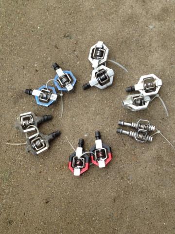 A collection of bicycle pedal cleats arranged in a circular pattern on a sandy surface. The cleats are of various colors, including blue, red, and silver, showcasing different designs. Sheltowee Trace - Laurel Lake Trail mountain bike trail.