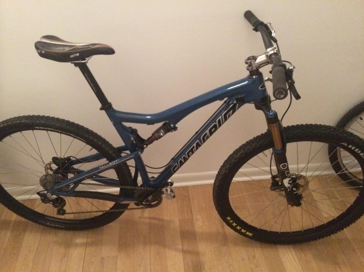 Santa Cruz Tallboy Carbon: A blue mountain bike with a sleek frame and knobby tires, positioned against a wall. The bike features a black seat, front suspension forks, and a visible gear system, sitting on a wooden floor.