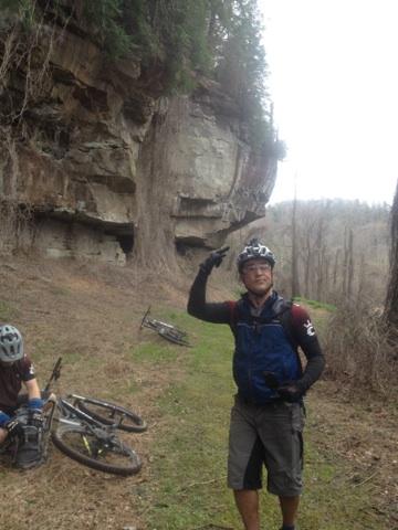 A mountain biker standing on a trail beside a rocky cliff, gesturing with one hand. In the background, another biker is sitting next to a bicycle on the ground. The landscape is wooded and hilly, with trees and rocky formations visible. Sheltowee Trace - Laurel Lake Trail mountain bike trail.