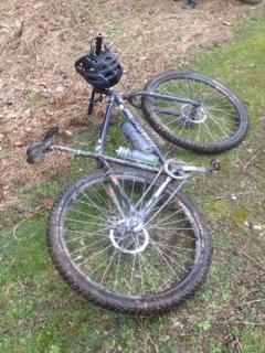A fallen mountain bike on the ground, partially on its side with a black helmet attached to the handlebars. The bike is covered in mud, indicating outdoor use, and is situated on a grassy area surrounded by dry foliage. Sheltowee Trace - Laurel Lake Trail mountain bike trail.