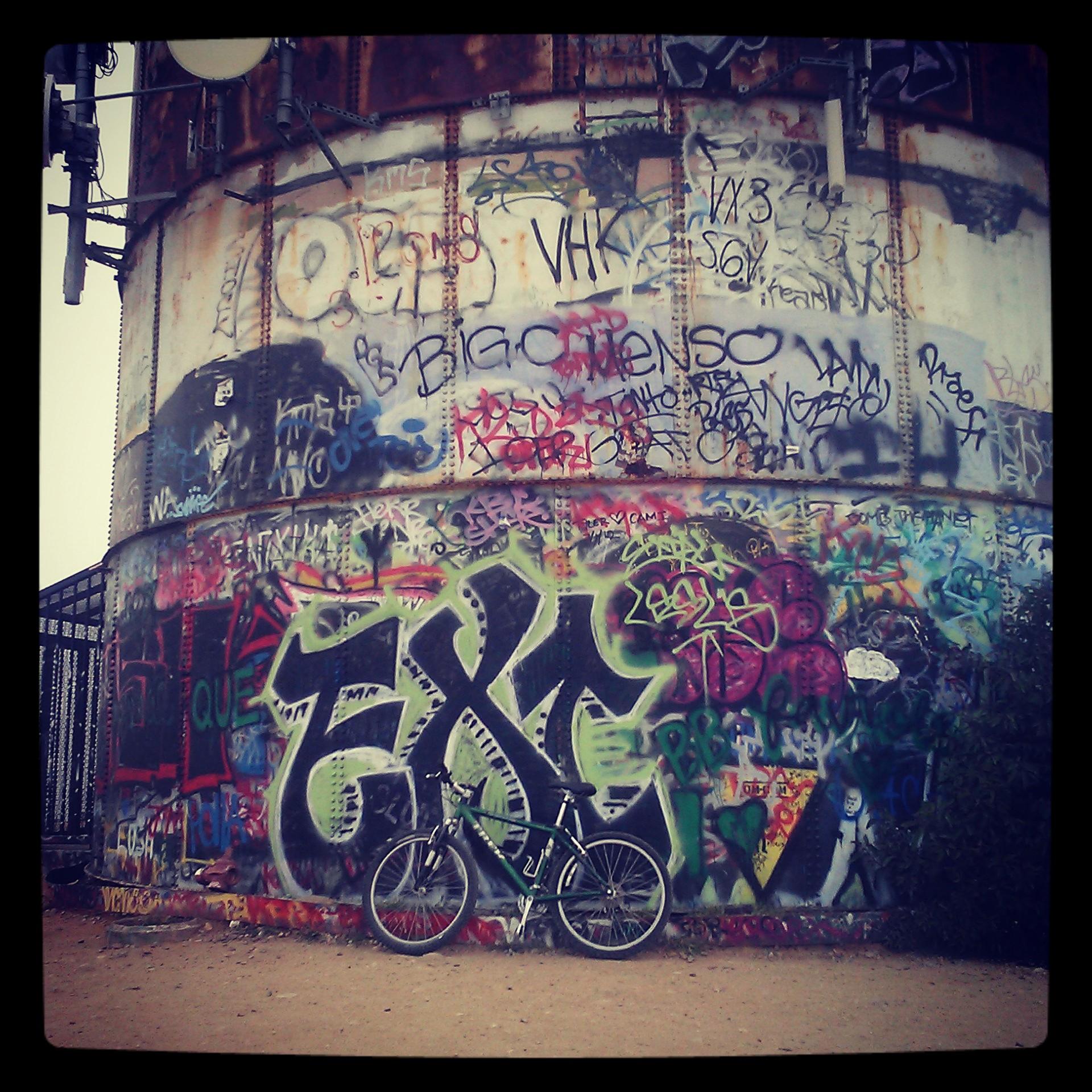 A graffiti-covered cylindrical structure with vibrant colorful designs and tags, featuring a black bicycle leaning against it. The surrounding area has gravel or dirt ground, and a few plants may be visible on the right side of the image. Turnbull Canyon mountain bike trail.