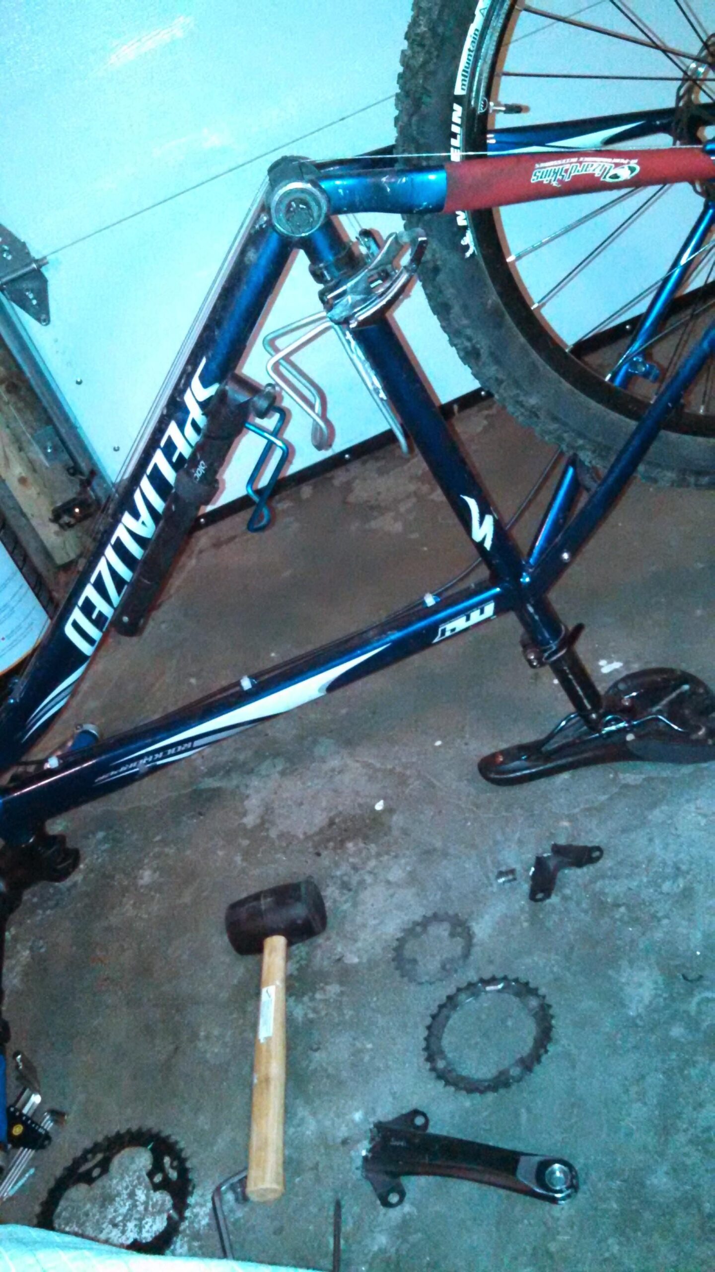 Specialized Rockhopper: A disassembled bicycle frame with a dark blue finish, lying on a concrete surface. The image includes various bike parts scattered around, such as a hammer, gears, and a crankset. The background features a garage setting with a white door partially visible.