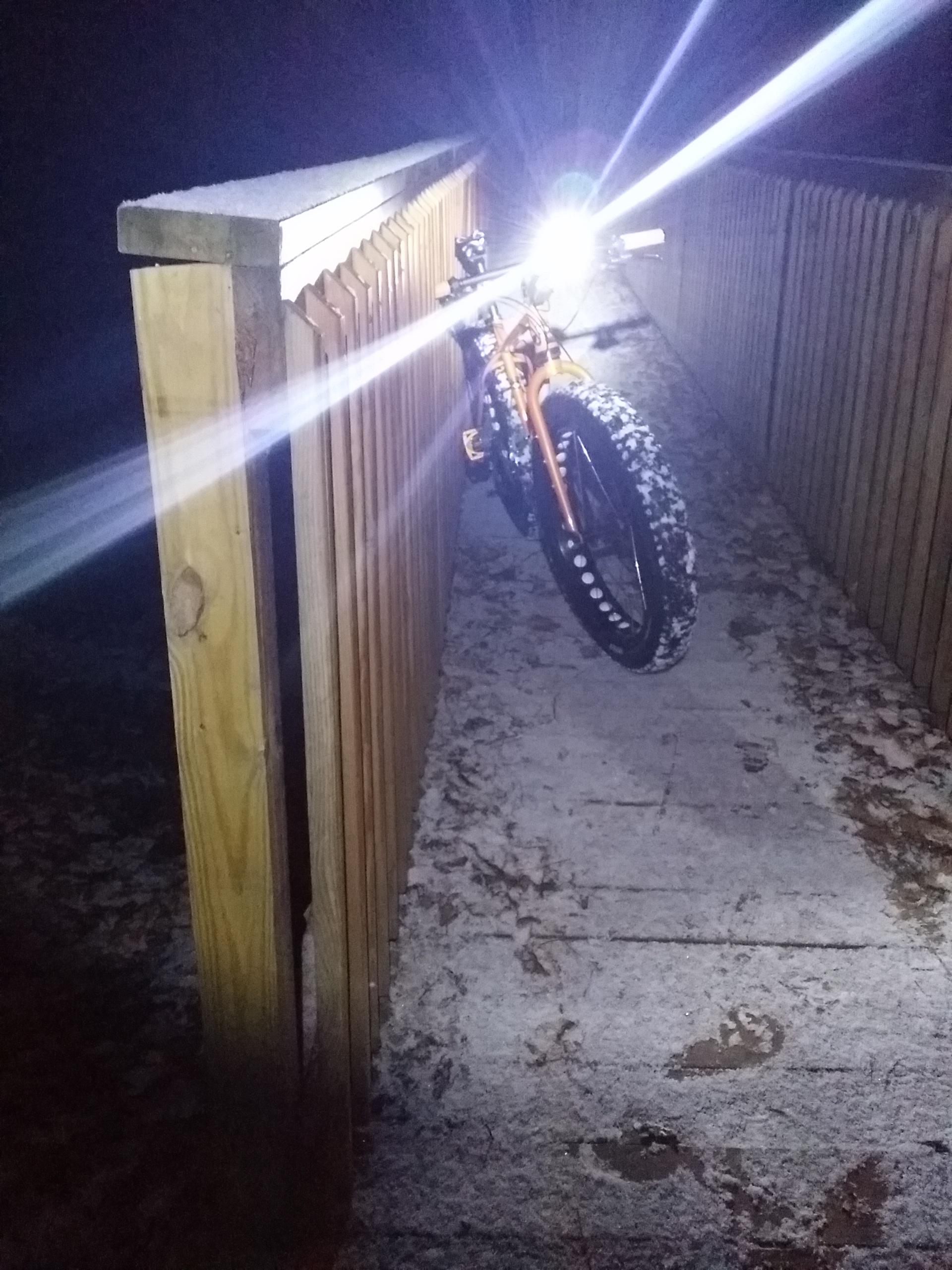 A fat bike leaning against a wooden railing on a snowy, dimly lit path at night, illuminated by the bike's front light, casting beams of light into the darkness. England Idlewild Mountain Biking Park mountain bike trail.