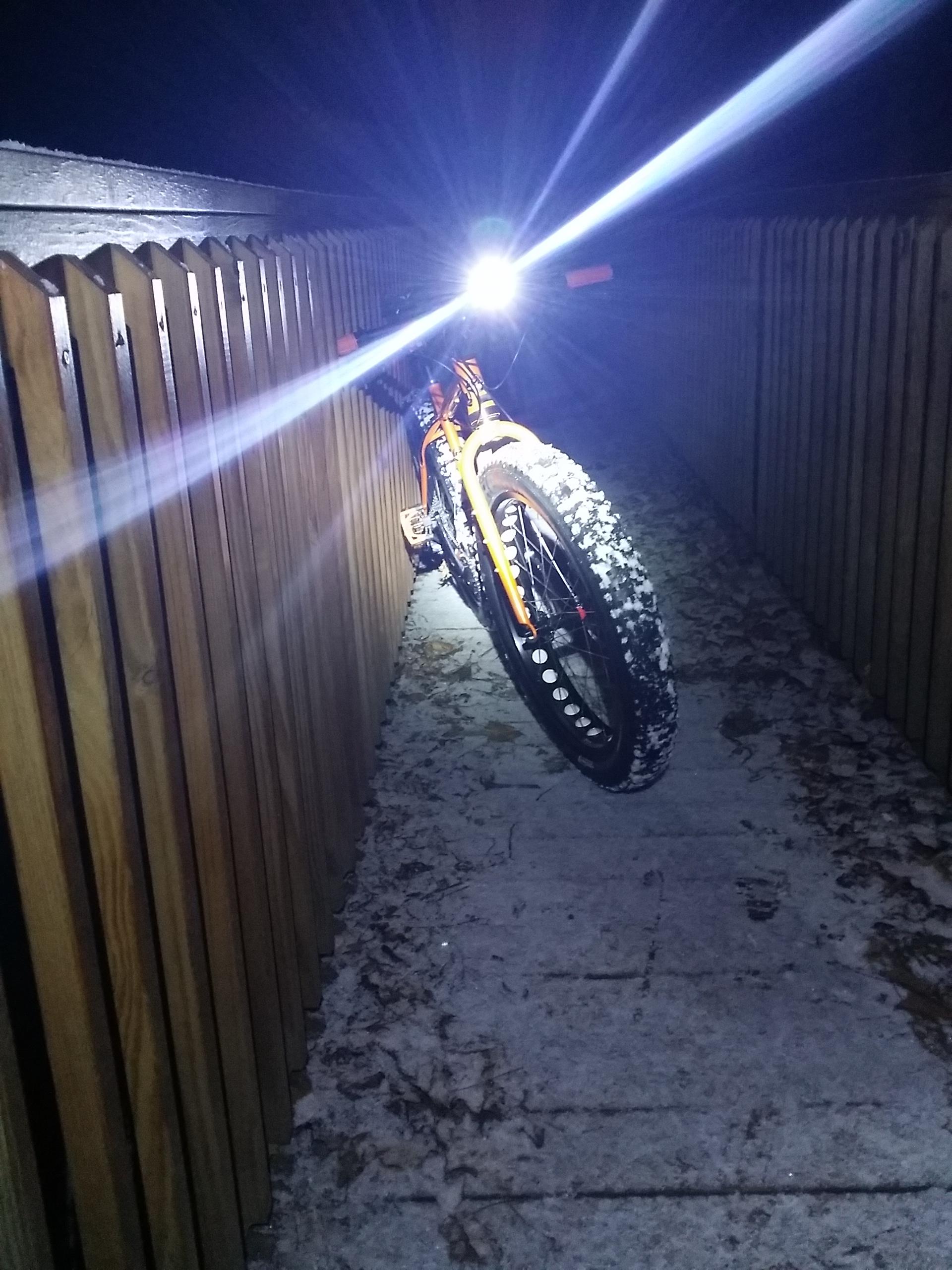 A brightly lit mountain bike with snow-covered tires rests against a wooden fence in a dimly lit pathway at night. England Idlewild Mountain Biking Park mountain bike trail.
