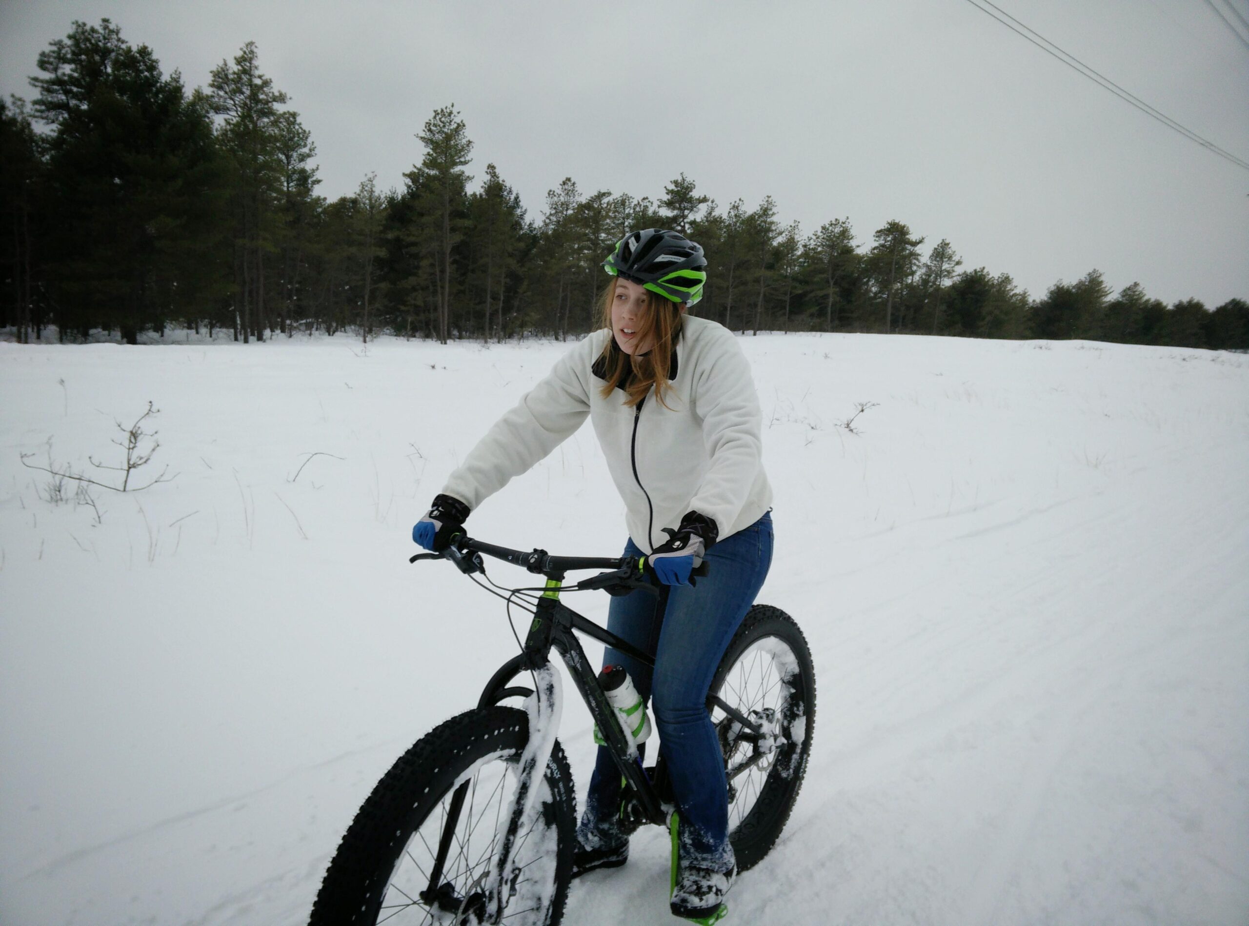 Trek Farley: A person riding a fat bike on a snowy trail surrounded by pine trees, wearing a black helmet, a white jacket, and blue gloves. The scene is set in a cold winter landscape with a cloudy sky.