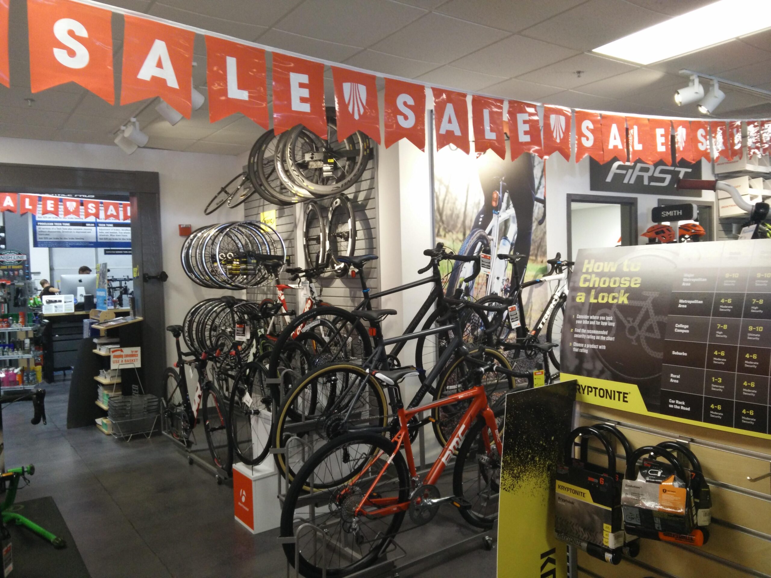 Bikes and bicycle wheels displayed in a shop with a prominent "SALE" banner overhead. A poster on the right provides tips on choosing a bike lock, while the background shows additional cycling accessories and a checkout area.