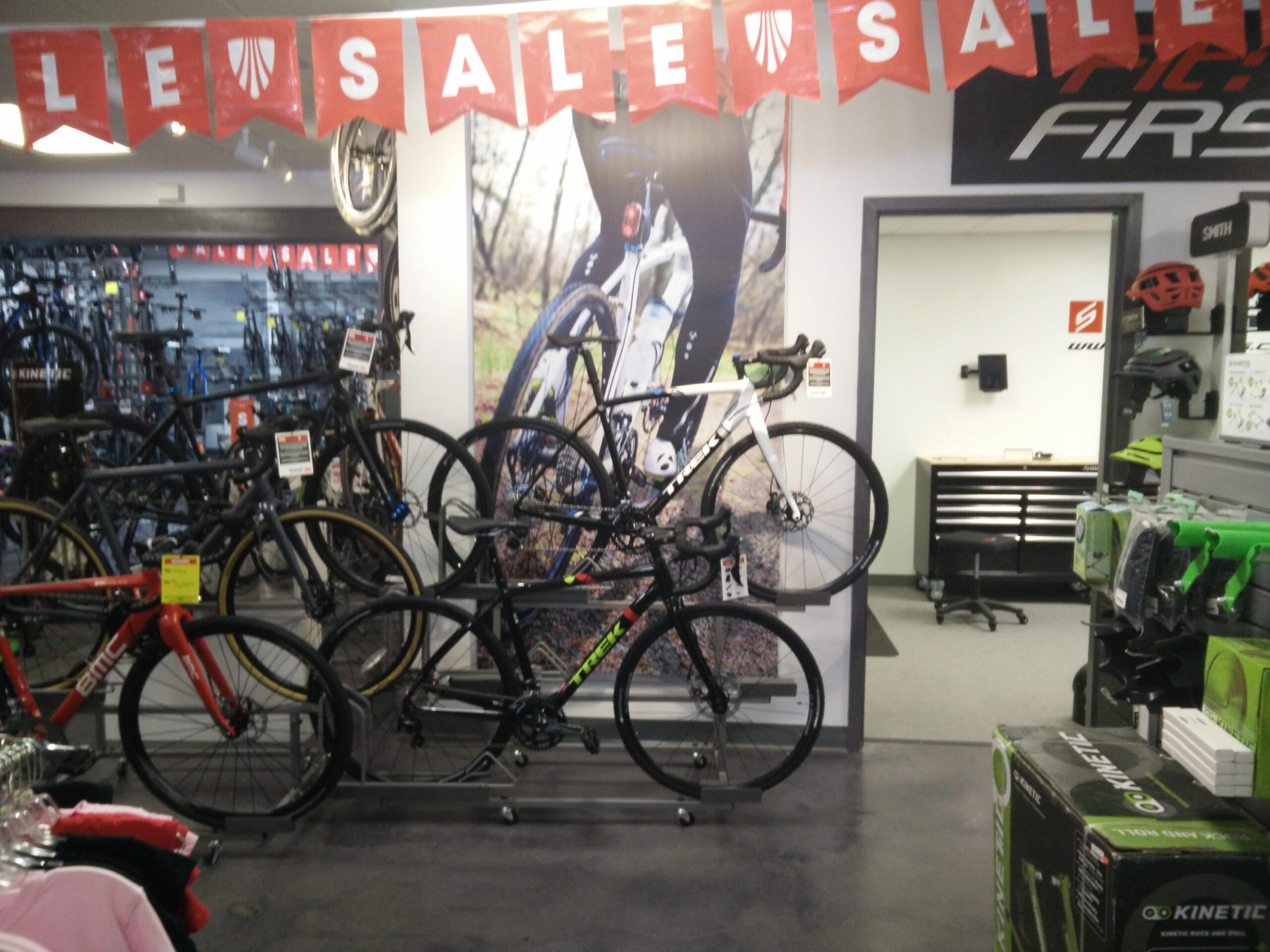 Alt text: An indoor bike shop featuring several bicycles on display, including black and red models. A large "SALE" banner hangs overhead. The shop has a variety of cycling gear and accessories visible, along with a sales counter in the background.