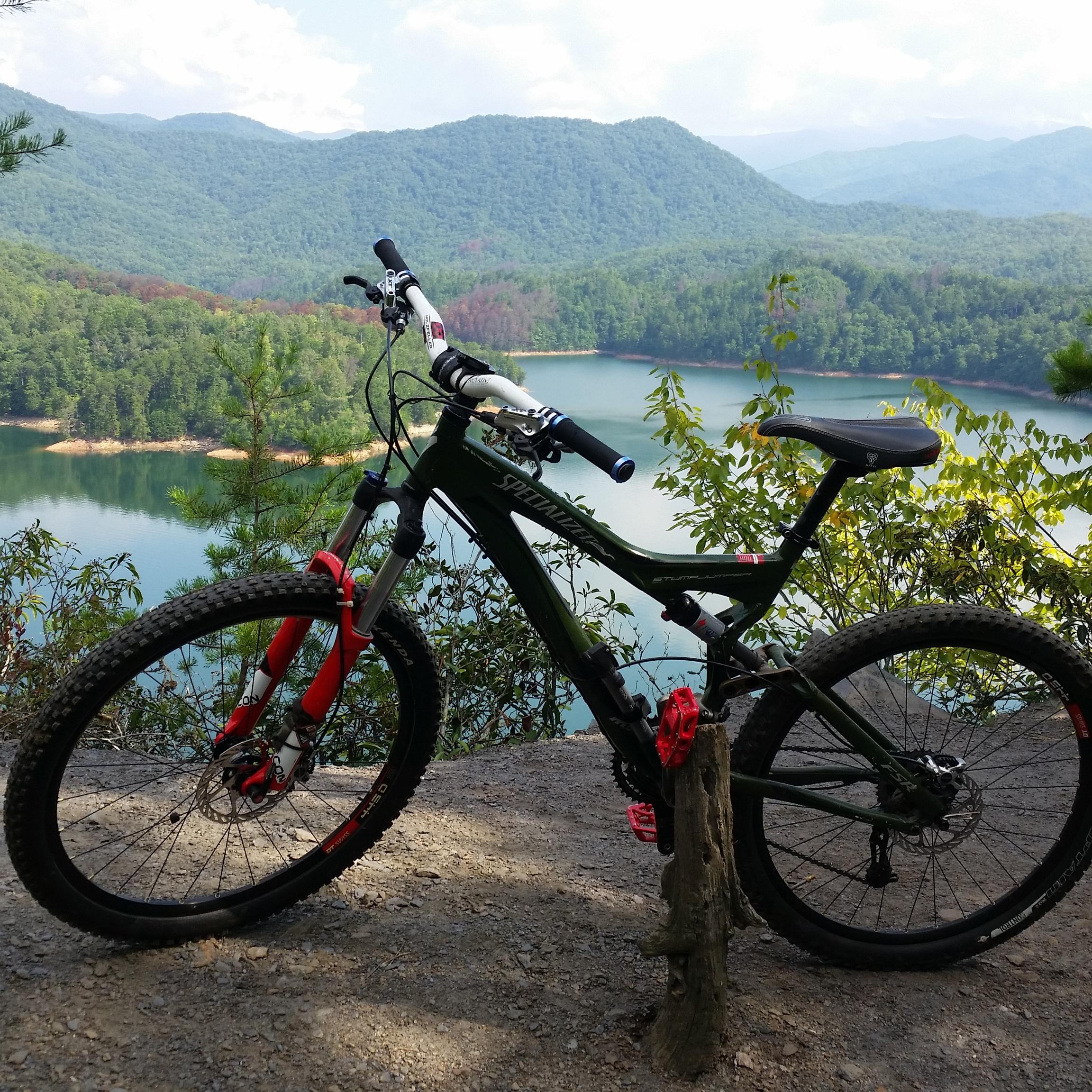 A mountain bike resting against a wooden post, with a stunning view of a river and mountains in the background. The lush green landscape reflects in the water, and the scene is brightened by a partly cloudy sky. Tsali Recreation Area mountain bike trail.