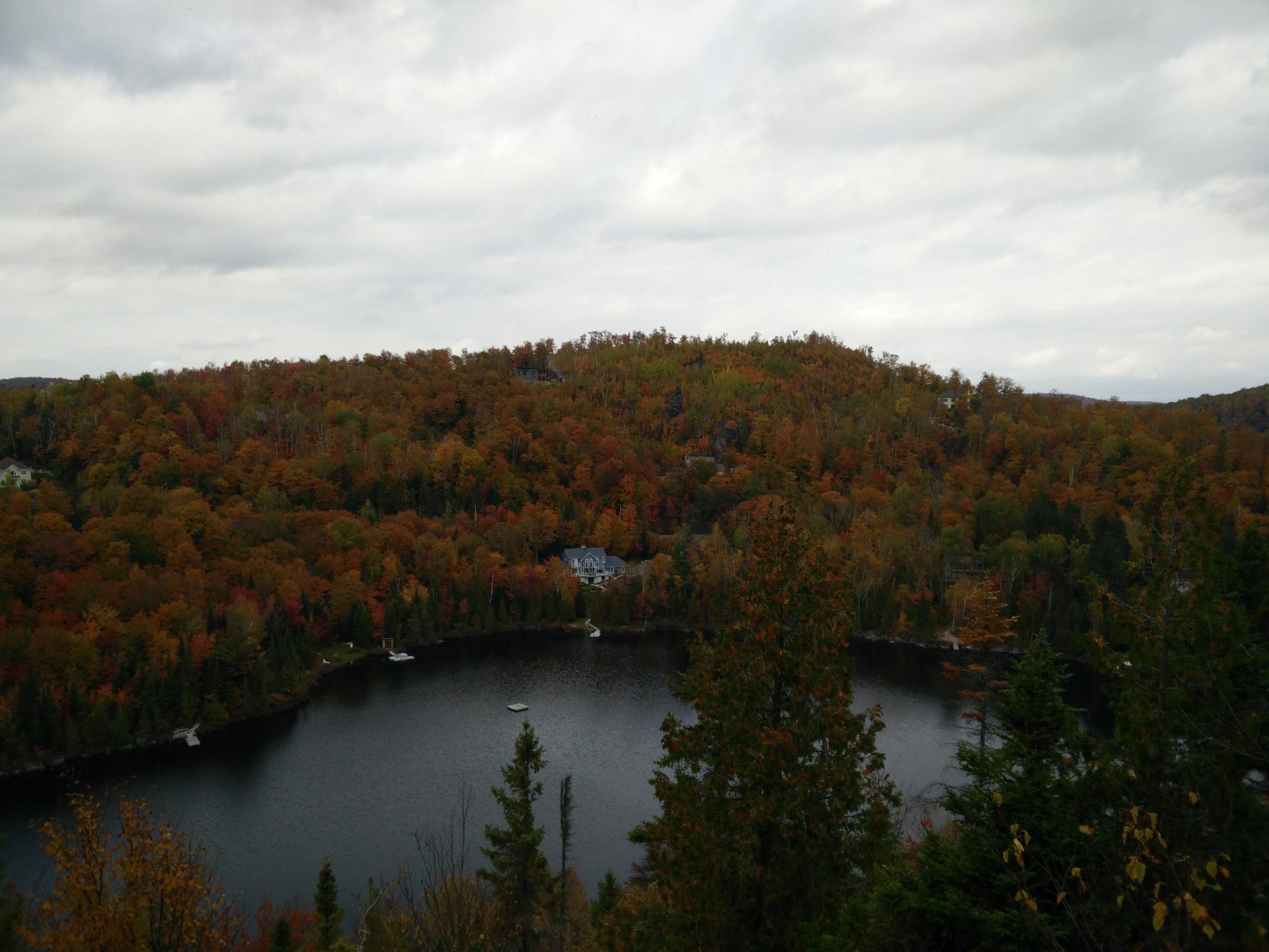 A scenic view of a tranquil lake surrounded by vibrant autumn foliage, with hills displaying a mix of orange, yellow, and green leaves under a cloudy sky. Several houses are visible near the water