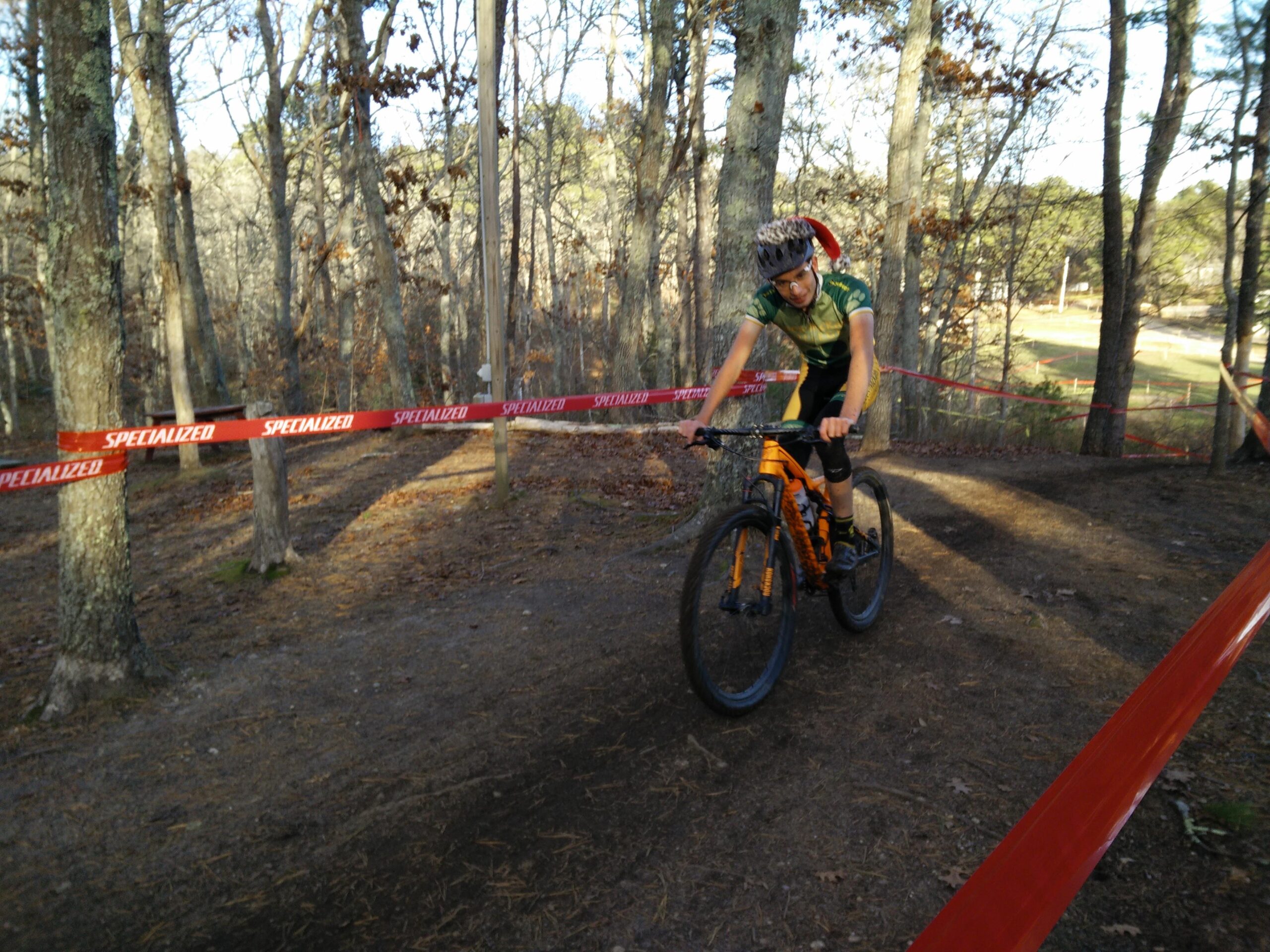 Specialized Epic: A mountain biker navigates a winding trail through a wooded area, wearing a green jersey and helmet. He is riding an orange bike, with a festive Santa hat visible on his head. Red tape marked "Specialized" is placed along the path, indicating a race course, while the sun filters through the trees, illuminating the scene.