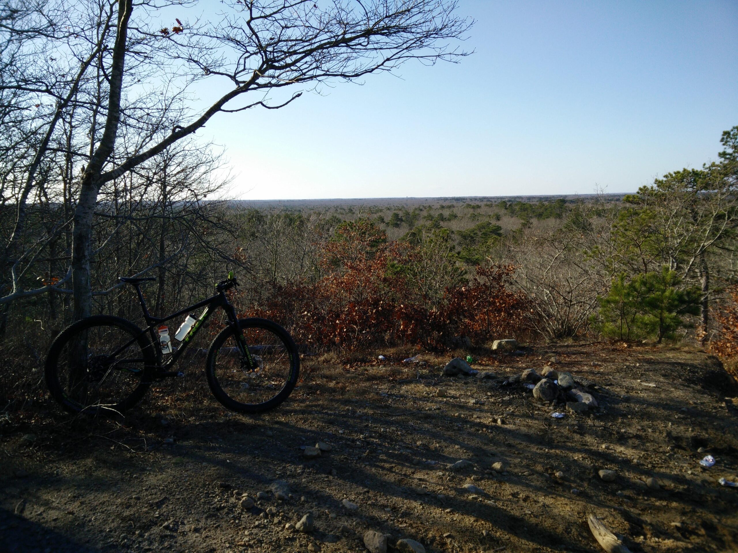 Trek Stache 9 29 Plus: A mountain bike rests near a rocky overlook, surrounded by bare trees and a sparse landscape. The horizon stretches out over a mixture of trees and open fields under a clear blue sky, with subtle shadows cast by the late afternoon sun.