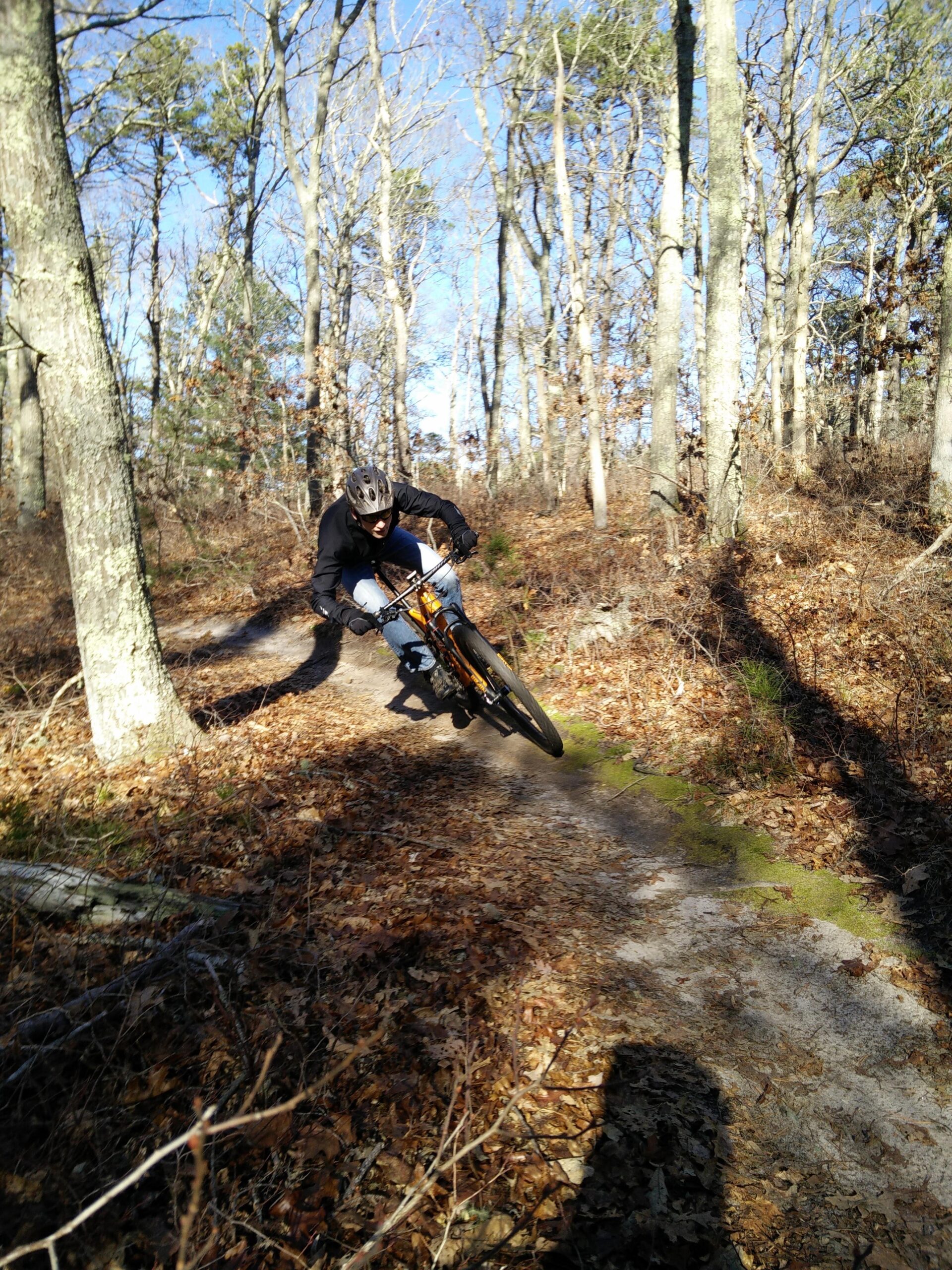 Specialized Epic: A mountain biker navigating a dirt trail through a wooded area with bare trees and fallen leaves, leaning into a turn. The scene is set on a sunny day with clear blue skies.