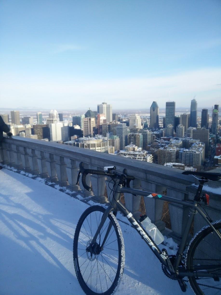 A bicycle standing on a snow-covered surface, overlooking a city skyline with tall buildings against a clear blue sky. The bicycle has thin tires, and in the background, a person can be seen enjoying the view. Parc du Mont-Royal mountain bike trail.