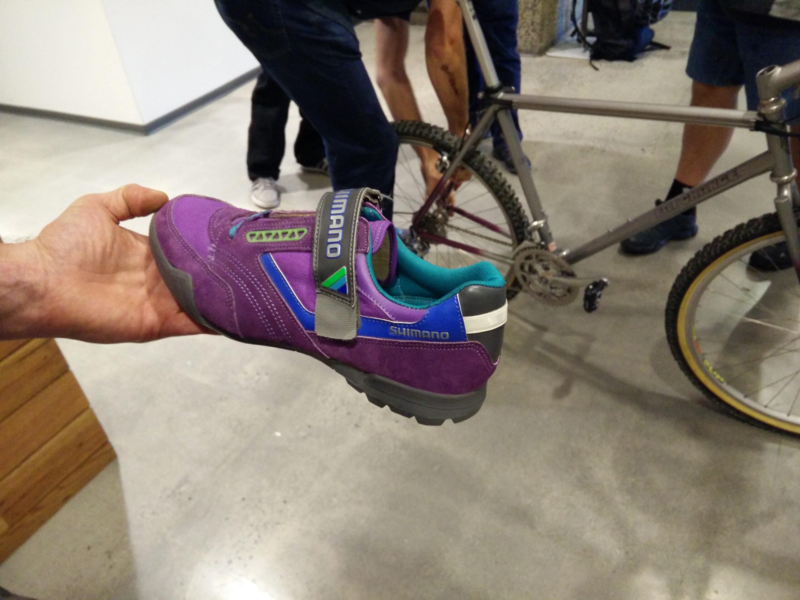 A hand holding a purple Shimano mountain bike shoe with a blue accent, set against a background of people working on a bicycle. The shoe features a Velcro strap and a rugged sole, indicative of cycling footwear.