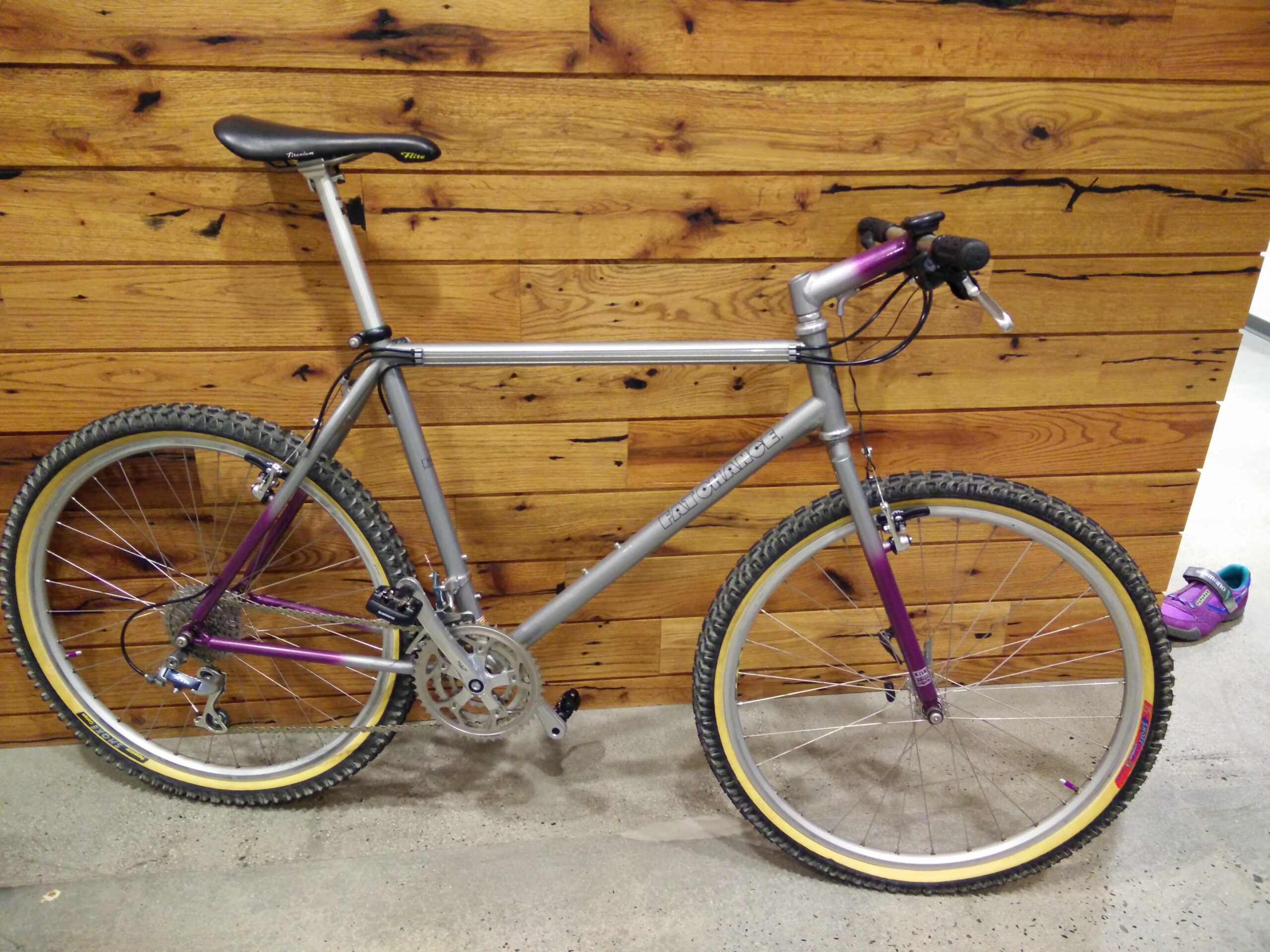 A silver and purple mountain bike is leaning against a wooden wall. The bike features thick tires designed for off-road use, and has a simple gear setup. A purple shoe is partially visible in the background on the floor.
