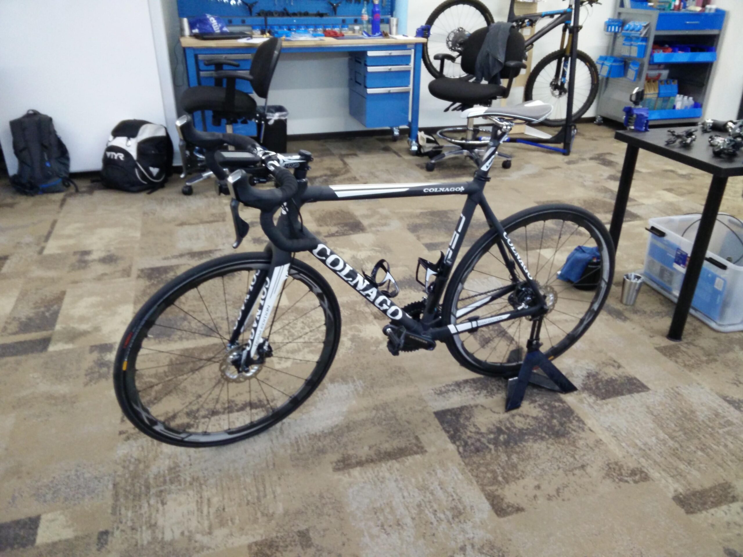 A black and white racing bicycle positioned on a repair stand, set in a workshop environment with tools and equipment in the background. Two backpacks are visible on the floor, along with a chair and a workbench filled with bike maintenance supplies.