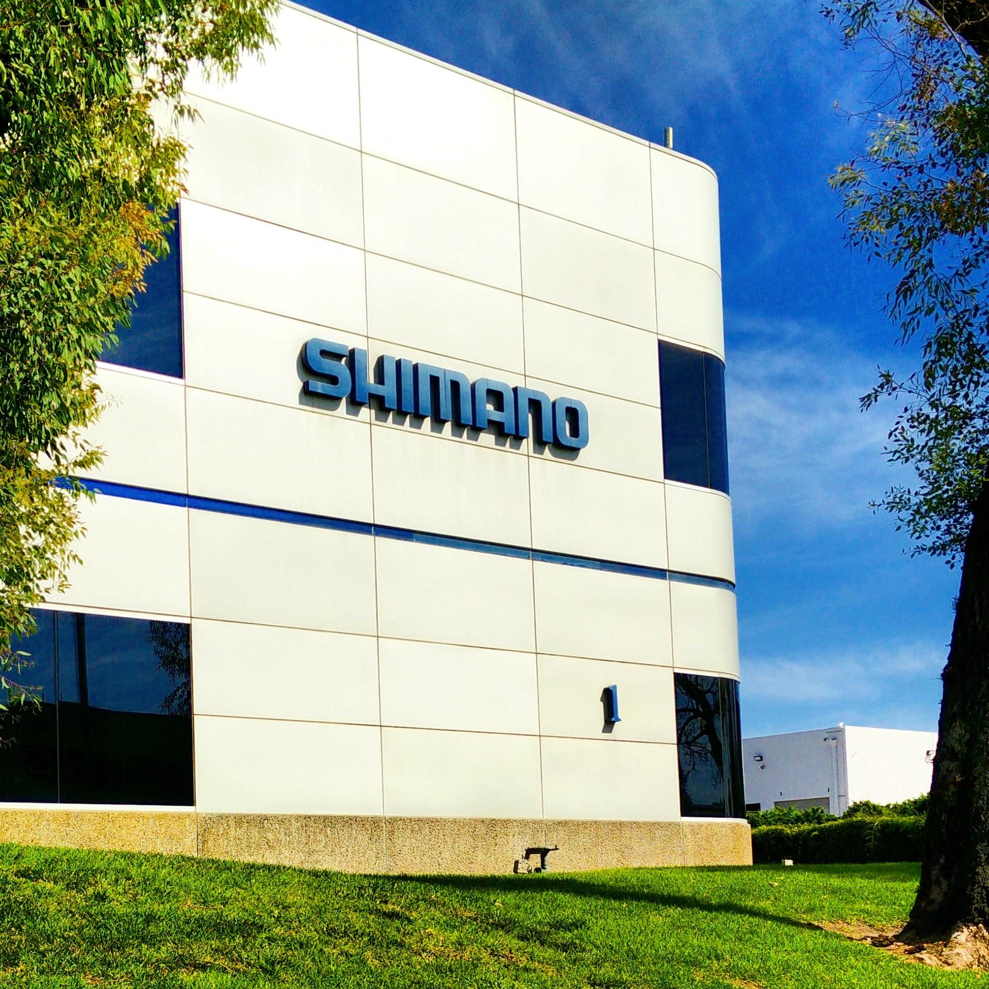 Modern building exterior featuring a sign for Shimano, with clear blue skies and landscaped greenery in the foreground.