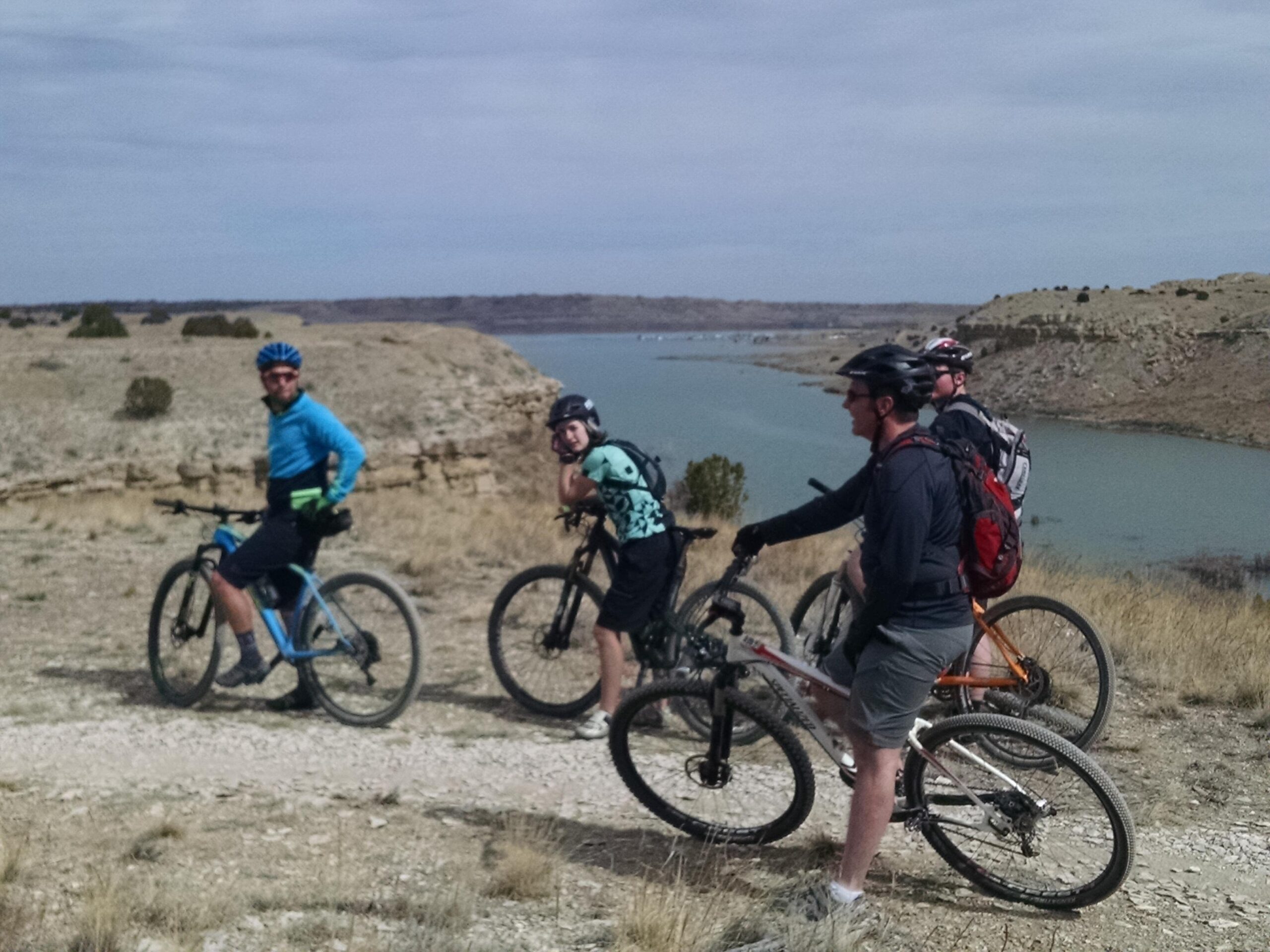 A group of four mountain bikers paused along a dirt trail overlooking a lake surrounded by rocky hills. They are dressed in casual cycling attire and helmets, with two riders smiling at the camera. The landscape features sparse vegetation and a cloudy sky, suggesting an outdoor adventure setting. South Shore Lake Pueblo mountain bike trail.
