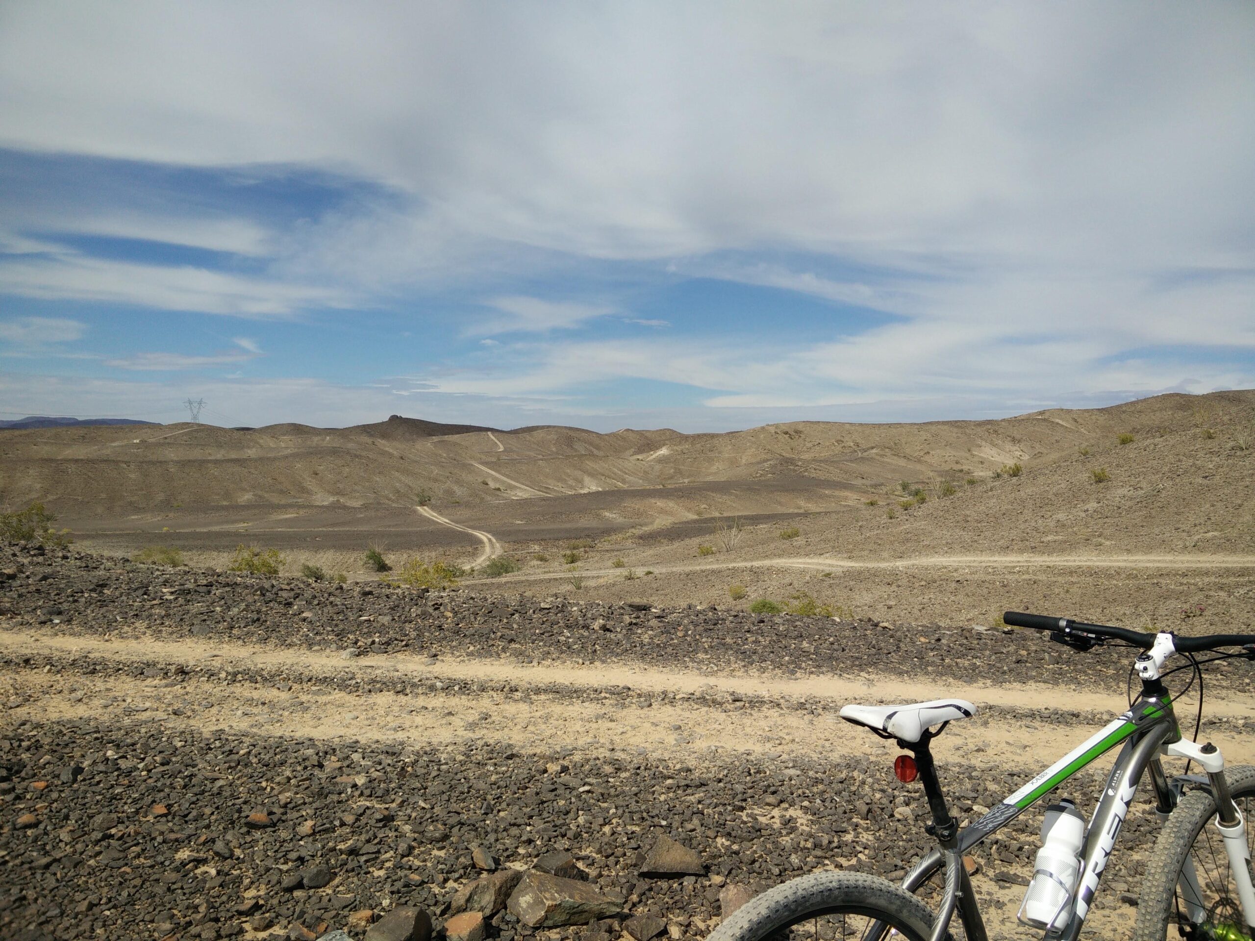 A mountain bike is parked on a rocky trail with a vast, dry landscape in the background. The scene features rolling hills and a clear blue sky with scattered clouds. Dusty paths wind through the terrain, suggesting a remote and rugged environment. Sugarloaf Peak mountain bike trail.