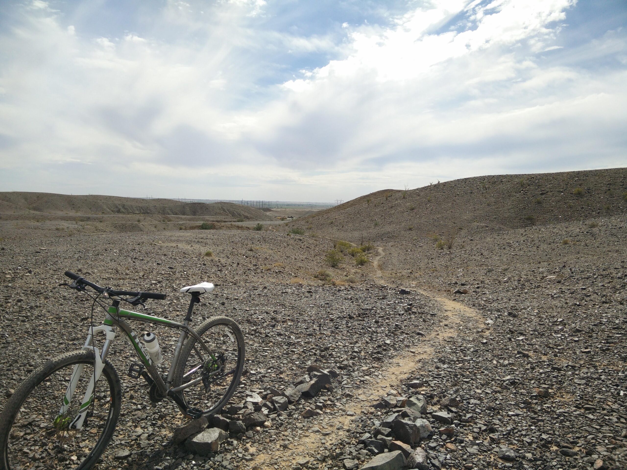 Mountain bike resting on a rocky trail with a scenic view of rolling hills under a partly cloudy sky. The landscape features a mix of rocky terrain and sparse vegetation, suggesting an outdoor cycling area. Sugarloaf Peak mountain bike trail.