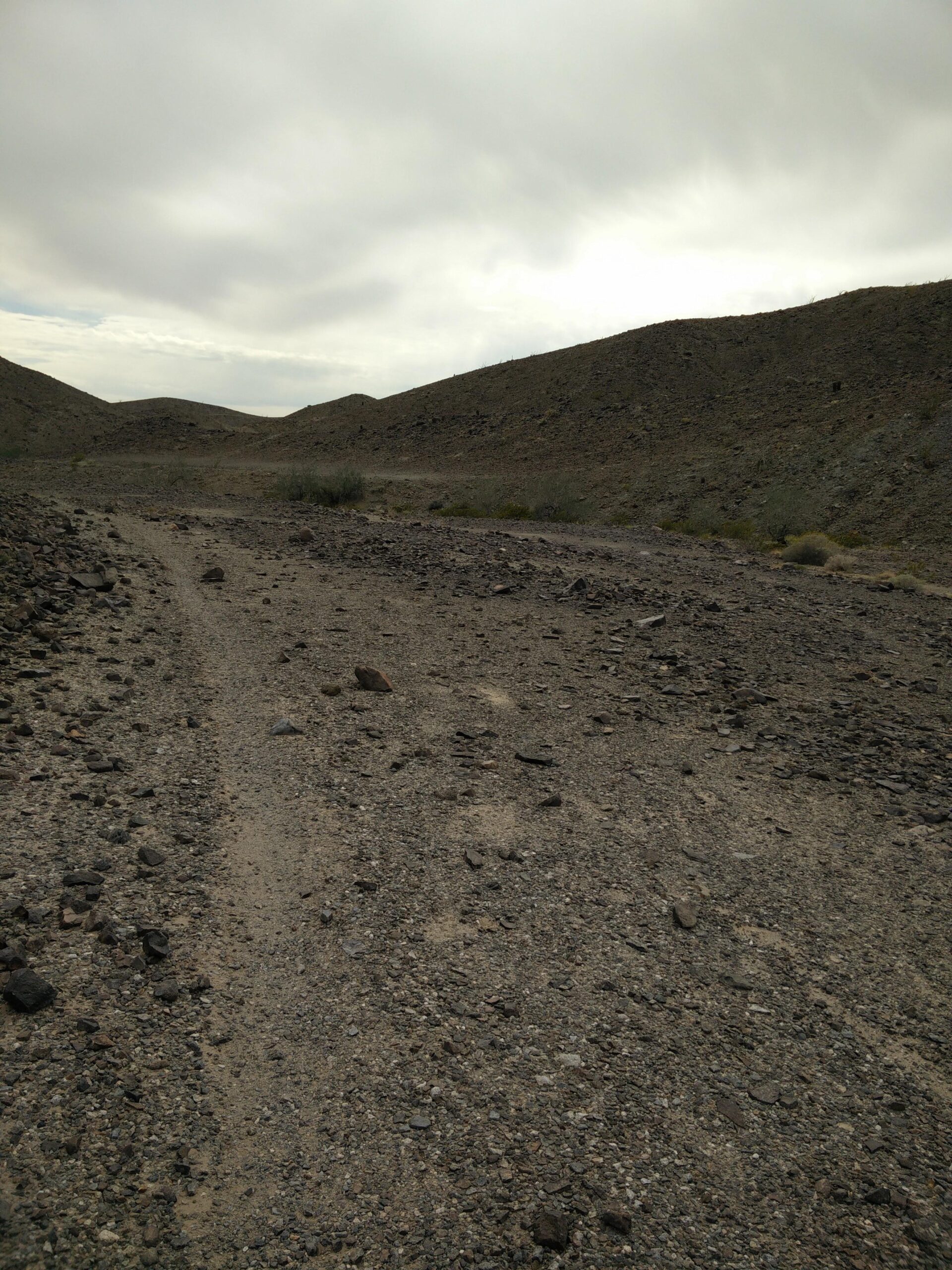 A gravel path winding through arid hills, with rocky terrain and sparse vegetation under a cloudy sky. Sugarloaf Peak mountain bike trail.
