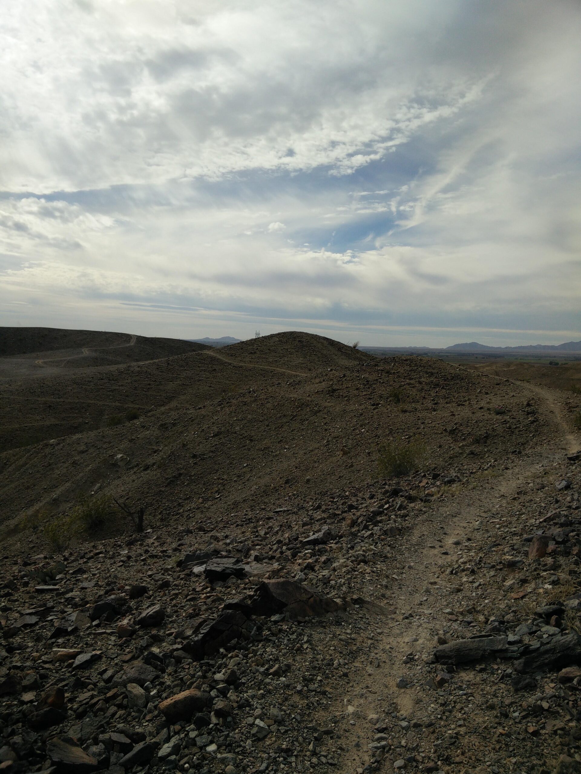 A rocky, desert landscape with rolling hills under a cloudy sky. A narrow dirt path winds through the foreground, surrounded by various stones and sparse vegetation. In the distance, more hills and mountains are visible against the horizon. Sugarloaf Peak mountain bike trail.
